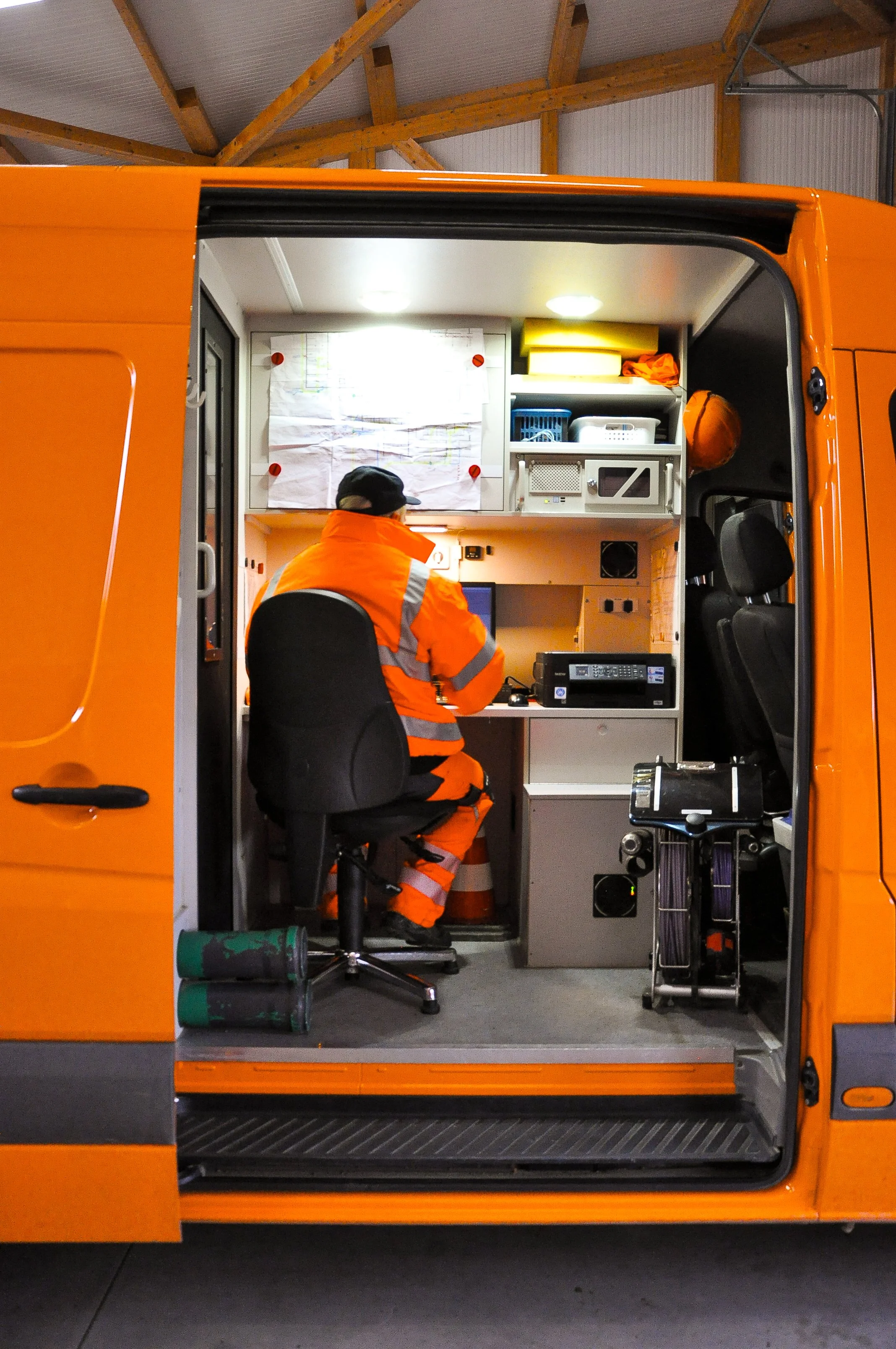 Ein Person in orangefarbener Warnweste und -hose sitzt in einem mobilen Einsatzwagen und arbeitet an einem Computer. Der Innenraum ist mit Geräten und Dokumenten ausgestattet.