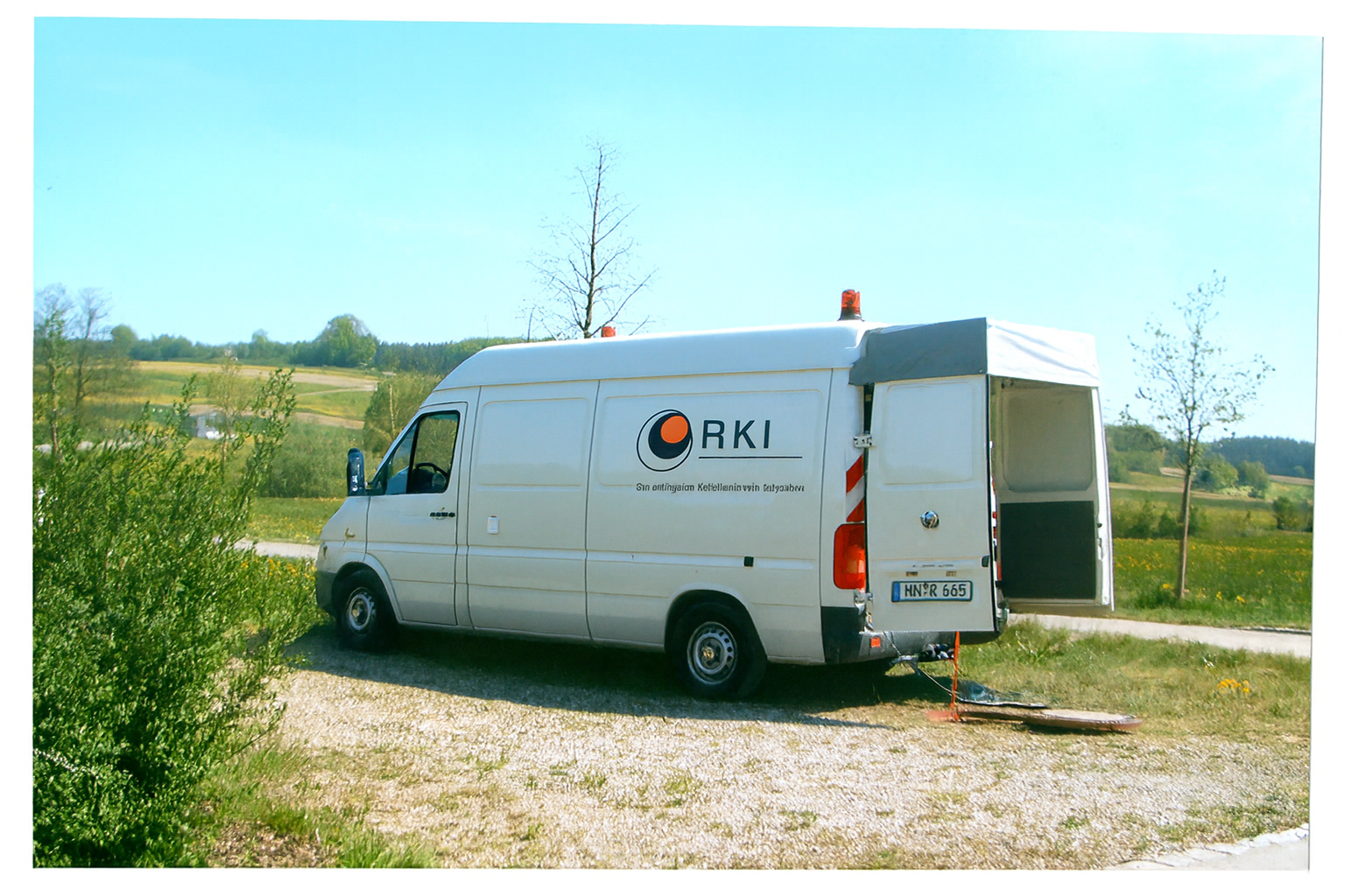 Ein weißer Dienstwagen mit dem Logo und Schriftzug von RKI steht auf einem Feld, der Hinterwagen ist offen, es ist eine ländliche Umgebung mit Bäumen und Hügeln im Hintergrund.