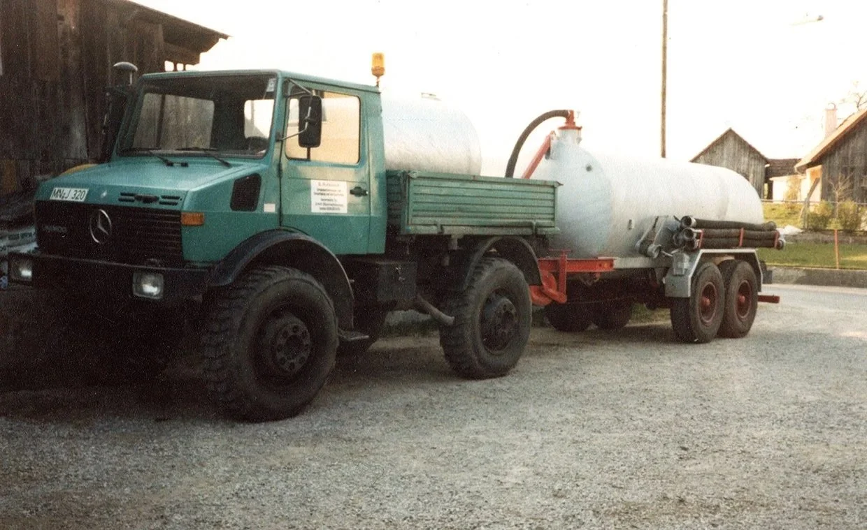 Ein gemischter Wasser- und Tankwagen auf einer Straße, umgeben von ländlichen Gebäuden und Bäumen.