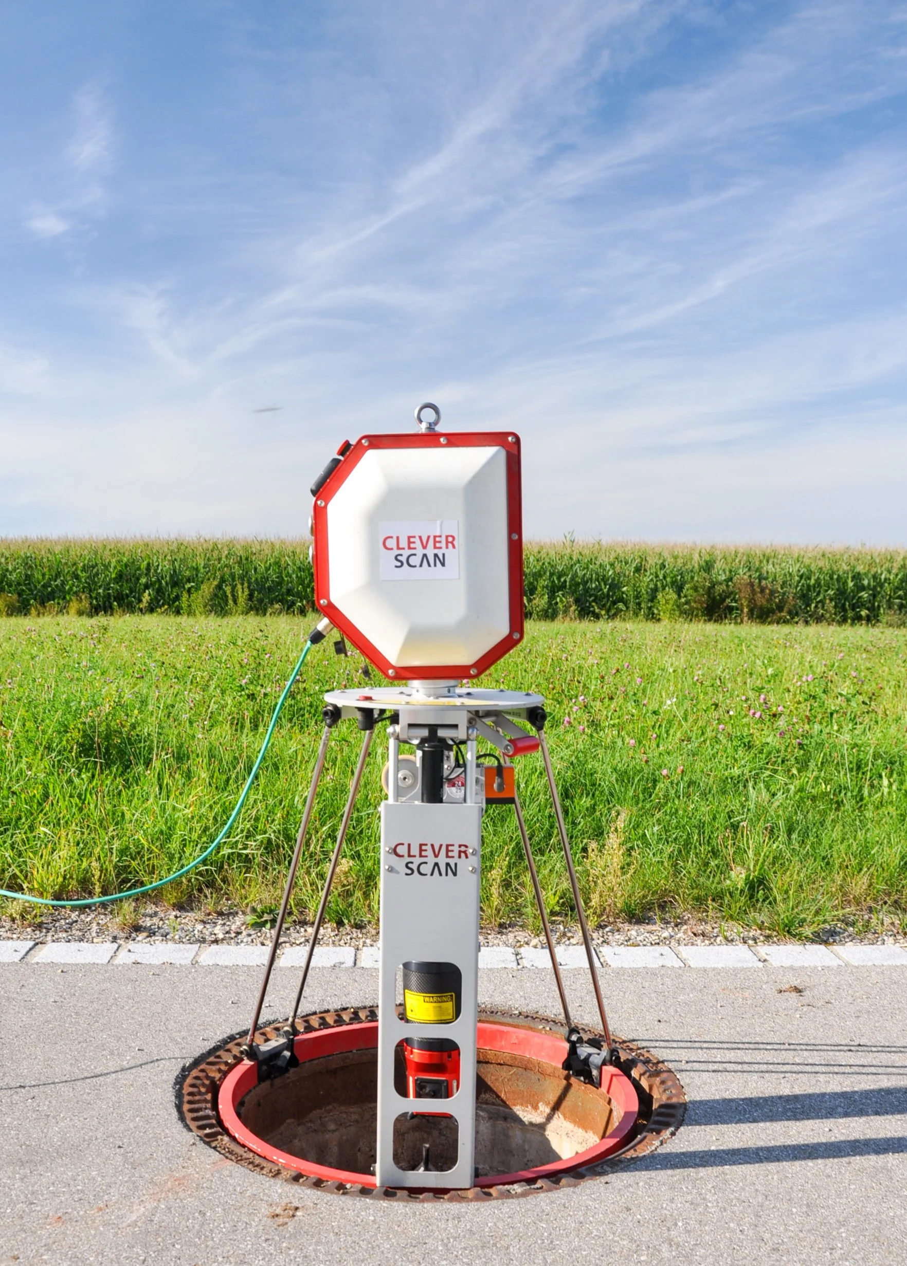 Eine Schachtkamera der Marke Clever Scan befindet sich auf einer Straße, mit einem offenen Schachtdecke davor, im Hintergrund grüne Wiese und blauer Himmel.