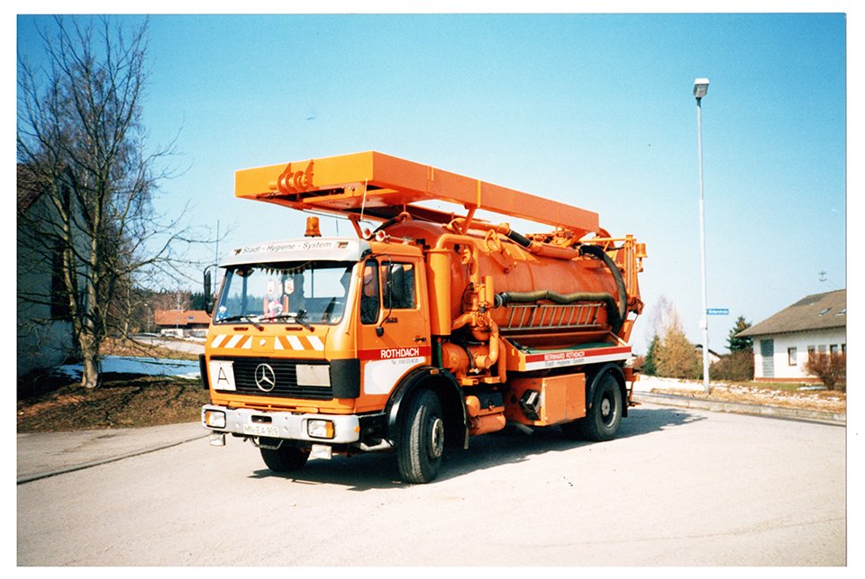 Ein orangefarbener Kanalspüler auf einem Straßengelände mit einem Haus und Bäumen im Hintergrund.