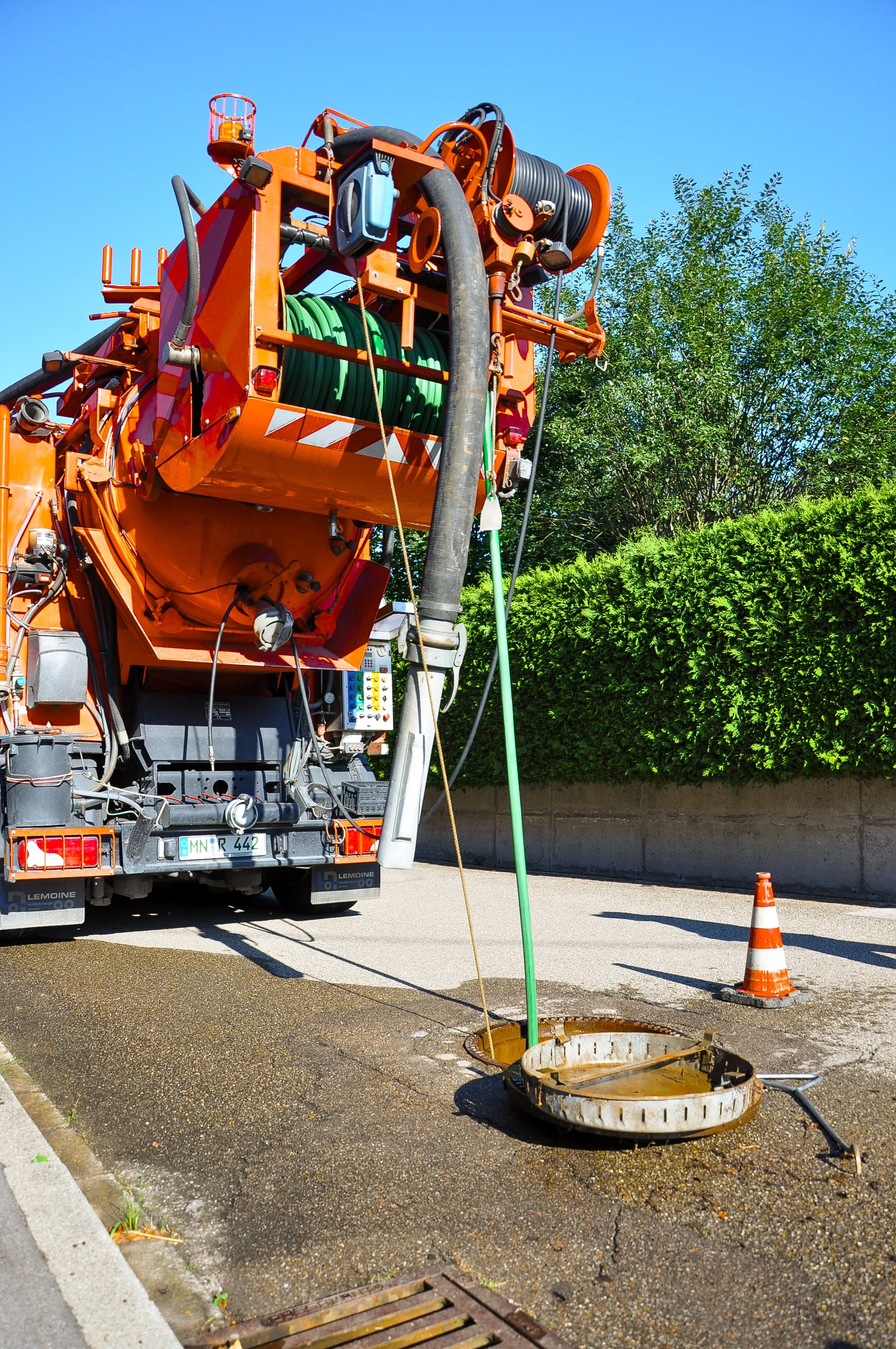 Ein orangefarbener Straßenreinigungswagen mit Schlauch und einem geöffneten Mannlochdeckel auf der Straße.
