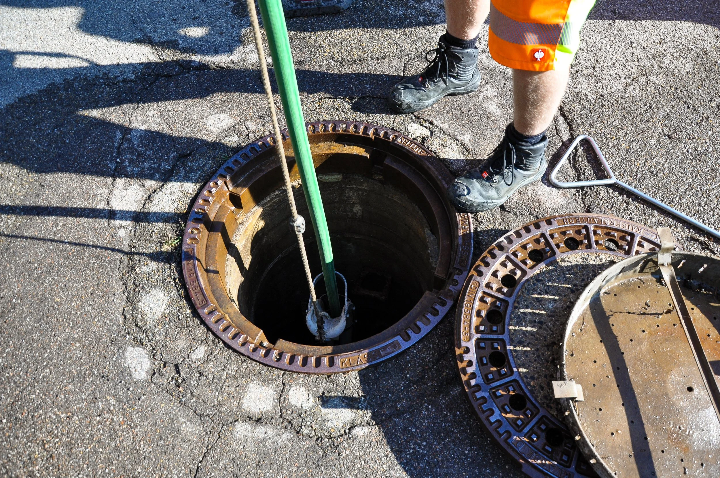 Mitarbeiter mit Sicherheitsstiefeln und orangefarbener Warnweste öffnet eine abgedeckte Straßenöffnung, um Zugang zu unterirdischen Versorgungssystemen zu ermöglichen.