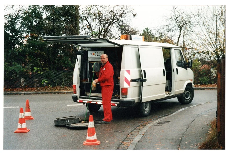 Ein Kanalreinigungs-Mitarbieter in roter Arbeitskleidung vor einem Kastenwagen. Der Wagen ist neben einem Fahrradständer auf einer Straße mit Bäumen im Hintergrund geparkt, einige Verkehrskegel sind aufgestellt.