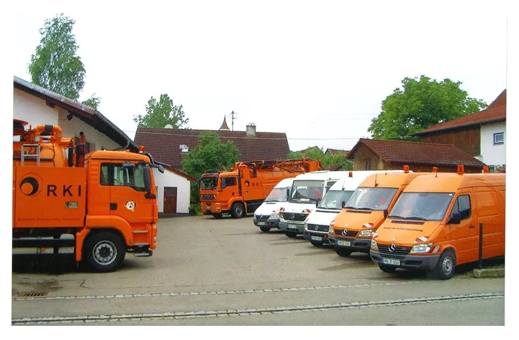 Parkplatz mit mehreren orangefarbenen RKI-Reinigungsfahrzeugen und weißen Lieferwagen, umgeben von Häusern mit roten Dächern und grünen Bäumen.