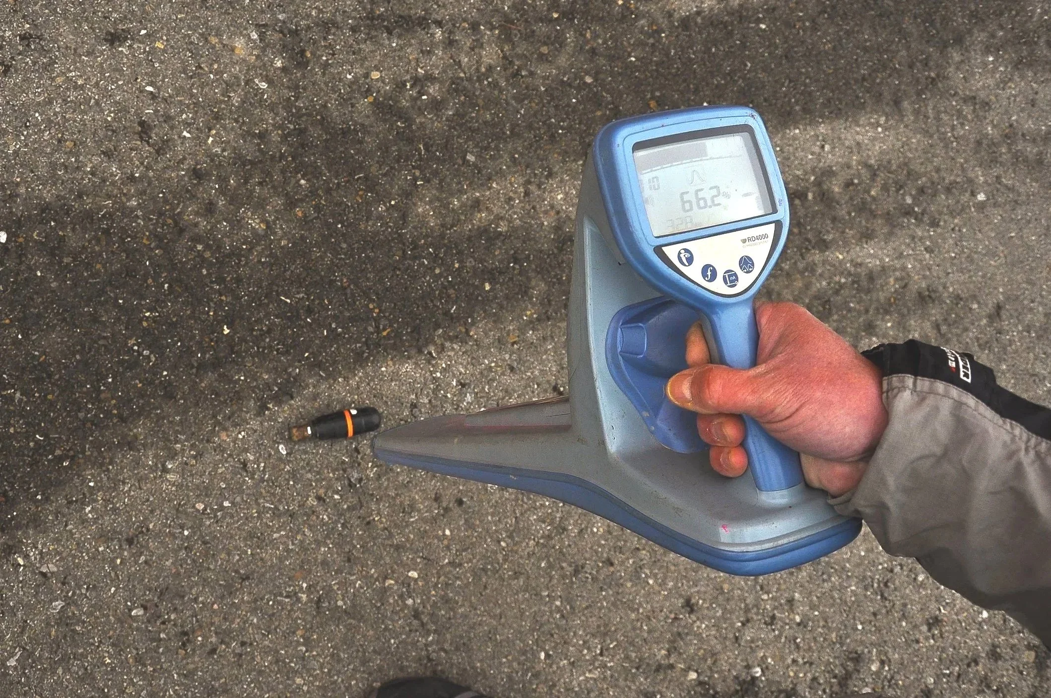 Hand hält ein digitales Ortungsgerät, das die Kanal Sonde die am Boden liegt erkennt.