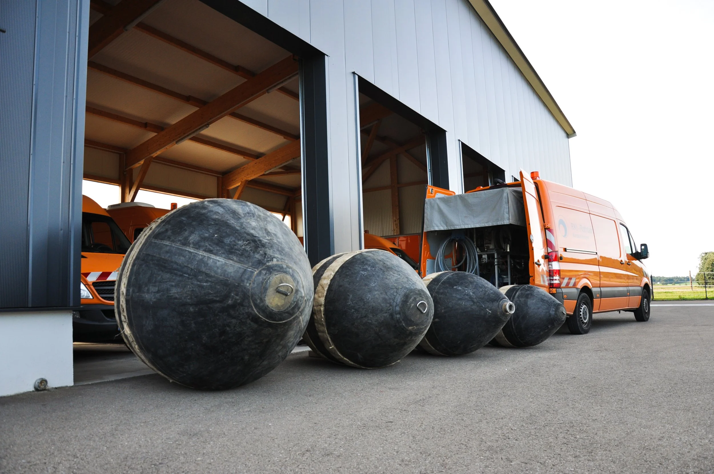 Ein Lagerraum mit orangefarbenen Arbeitsfahrzeugen, vor dem eine Reihe von großen, schwarzen Blasen für Kanal Dichtheitsprüfungen liegen.