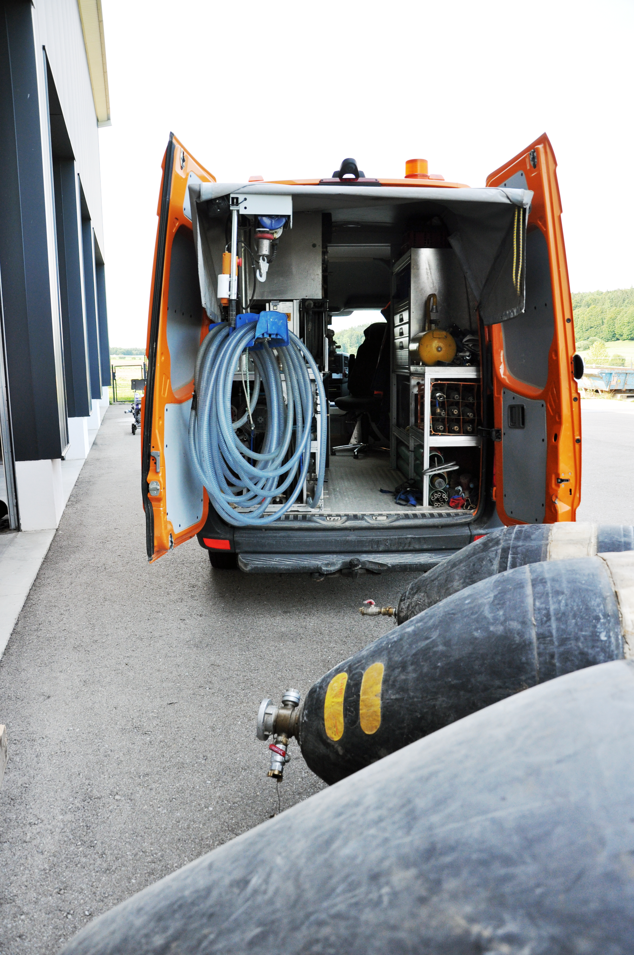 Ein orangefarbener Werkstattwagen mit geöffneten Türen, gefüllt mit Werkzeug und Ausrüstung, im Hintergrund eine Landschaft mit Wäldern und Feldern, vor dem Wagen liegen Blasen zur Kanal Dichtheitsprüfung.