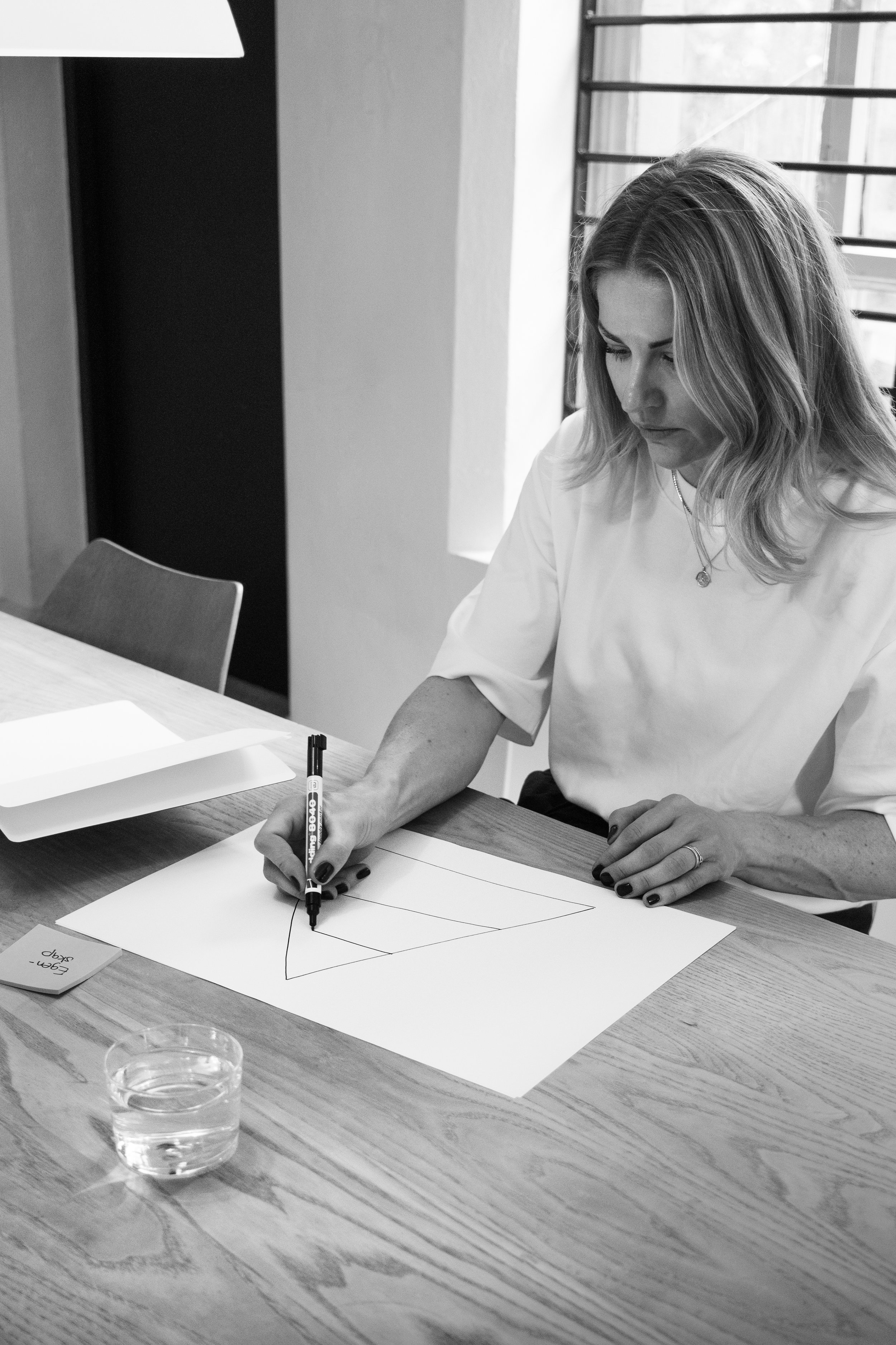 Marie in Bravuur with long hair and a necklace is sitting at a wooden table drawing geometric shapes on white paper with a black marker. There are papers, a pen, and a glass of water on the table, and a window with vertical blinds in the background.