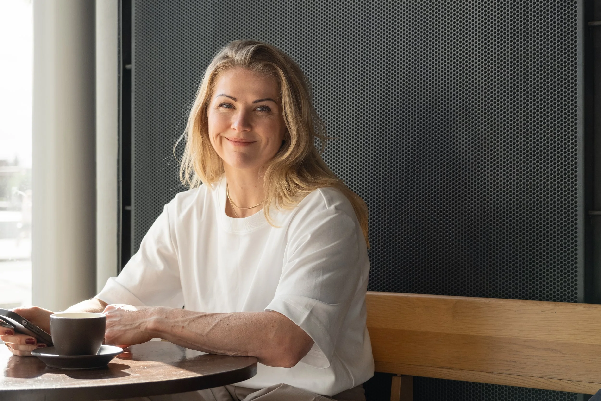 A woman with blonde hair sitting at a wooden table with a cup of coffee and a smartphone, smiling in a bright room with large windows.