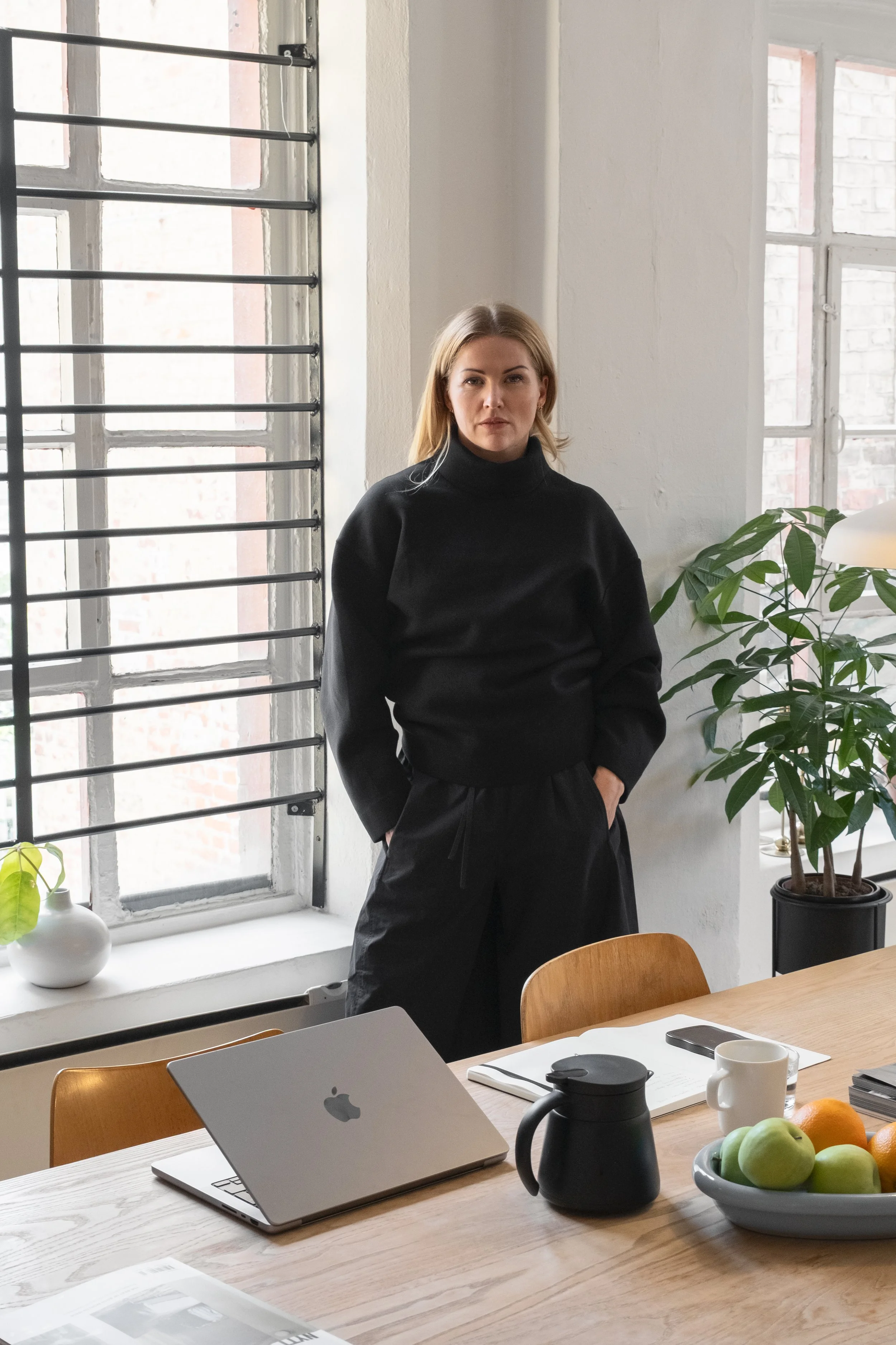 A woman in a black turtleneck standing next to a window with white walls and blinds. There is a potted plant on the windowsill to her right, and a table with a laptop, fruit, a mug, a thermos, and papers is in the foreground.
