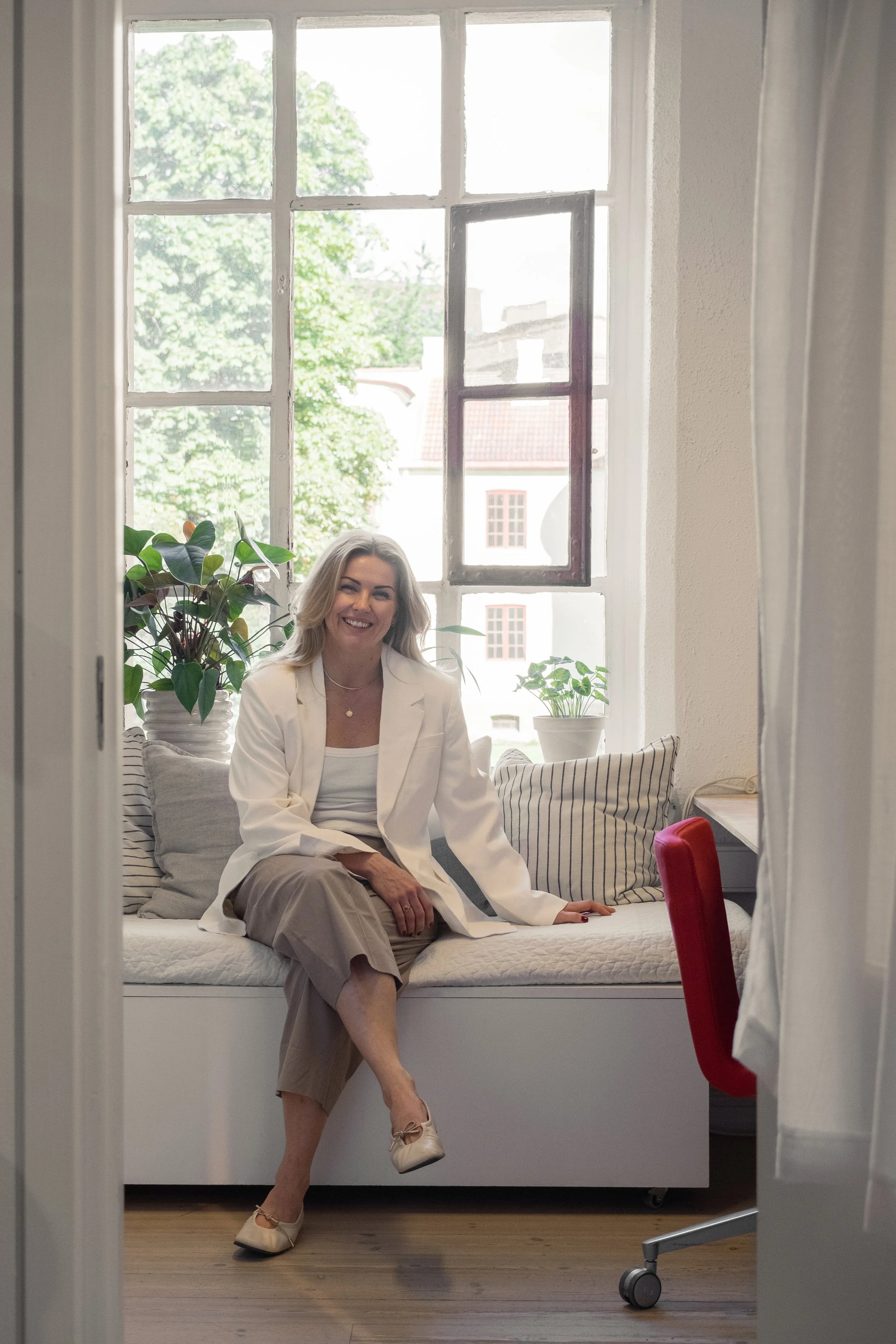A smiling woman with blonde hair, dressed in a white blazer and beige pants, sits on a bed decorated with striped pillows and green plants in white pots, in a room with large windows and natural light.