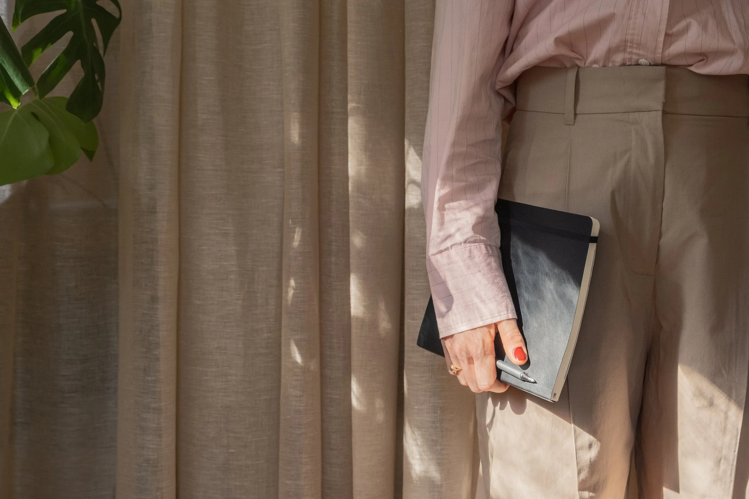 Person in pink striped shirt and beige pants holding a black notebook and a pen.