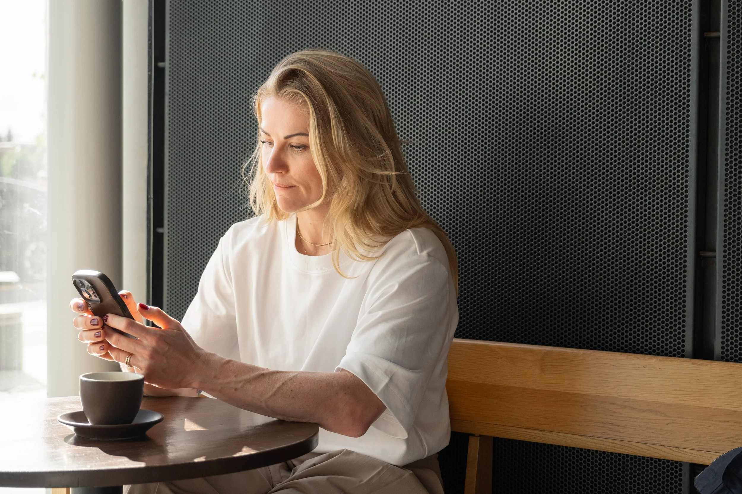 A woman with blonde hair sitting at a wooden table, looking at her smartphone with a cup of coffee in front of her, next to a large window.