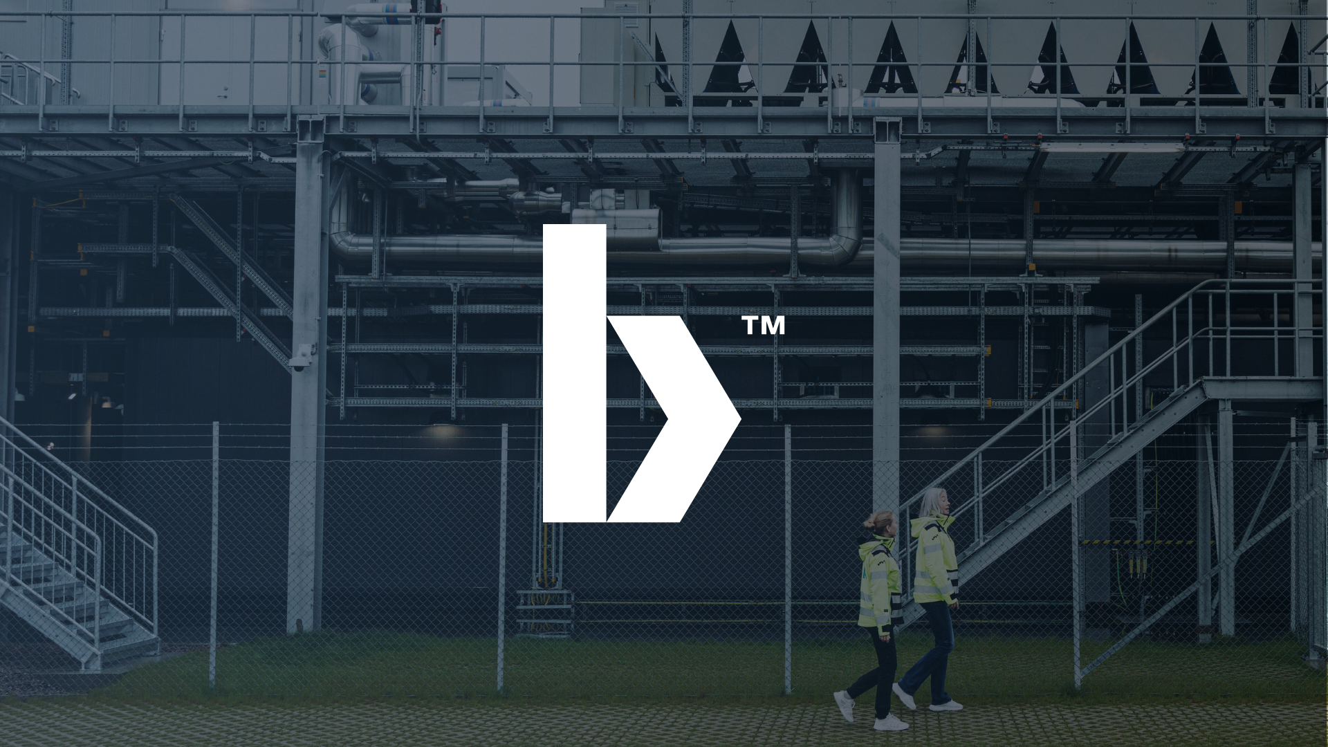 The image shows two women in high-visibility jackets walking along a chain-link fence outside an industrial facility with metal staircases, pipes, and structural beams. A large logo with a stylized arrow and the trademark symbol is overlaid on the center of the image.
