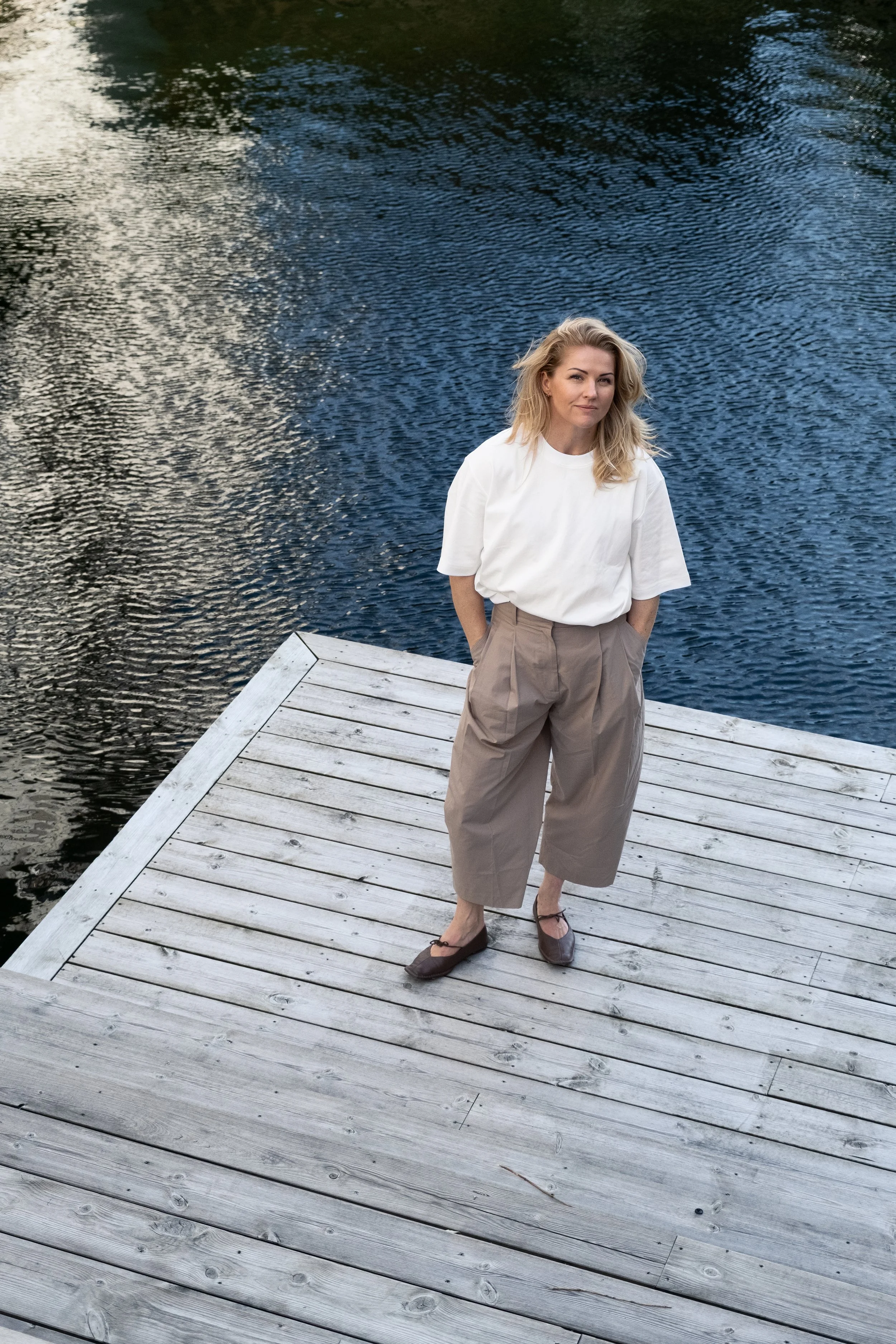 Marie in Bravuur standing on a wooden dock by a body of water, wearing a white top and beige wide-leg pants, with her hands in her pockets and looking at the camera.