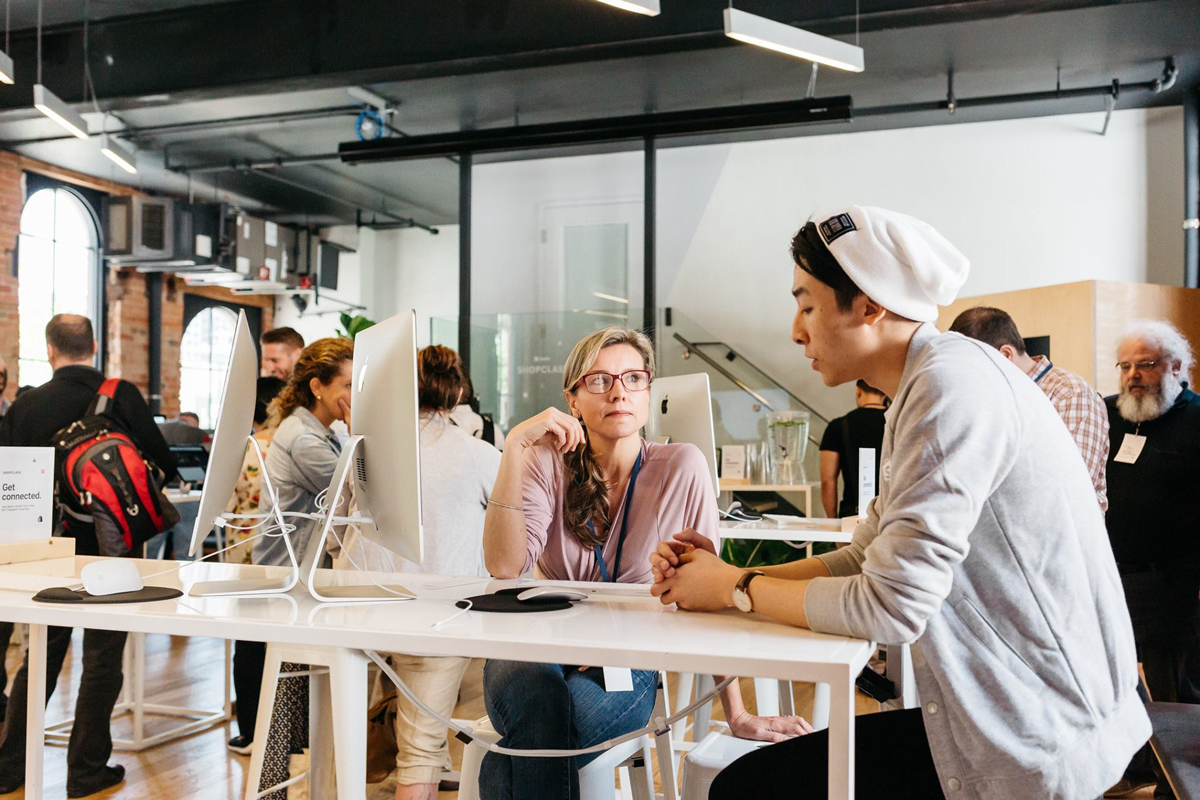 A woman with glasses and a purple shirt talking to a young man wearing a white hat and gray hoodie at a tech conference or coworking space with multiple people working on computers in the background.