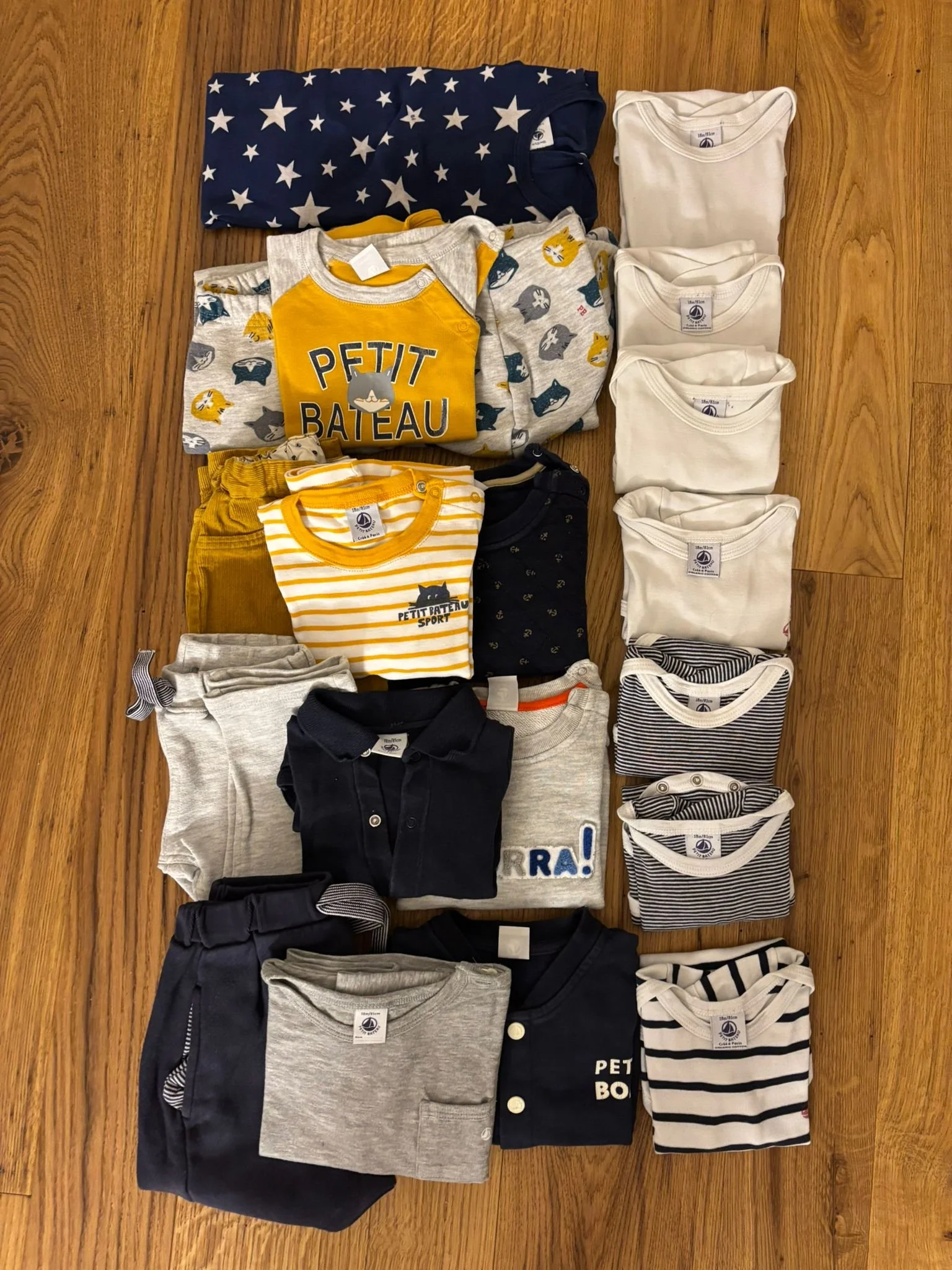 Collection of children's clothing including shirts, pants, and sweaters, neatly folded and arranged on a wooden floor.