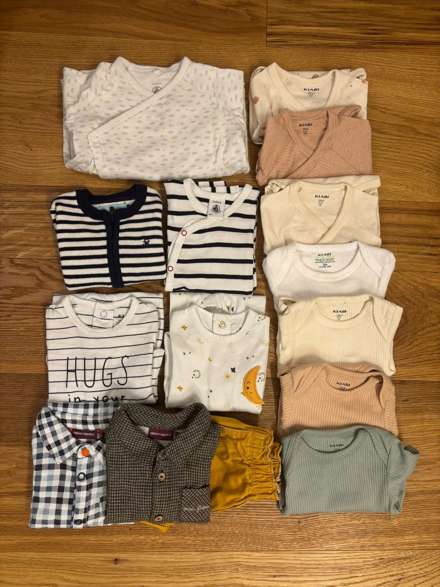 A collection of folded baby clothes on a wooden floor, including onesies, shirts, and pants in various colors and patterns.