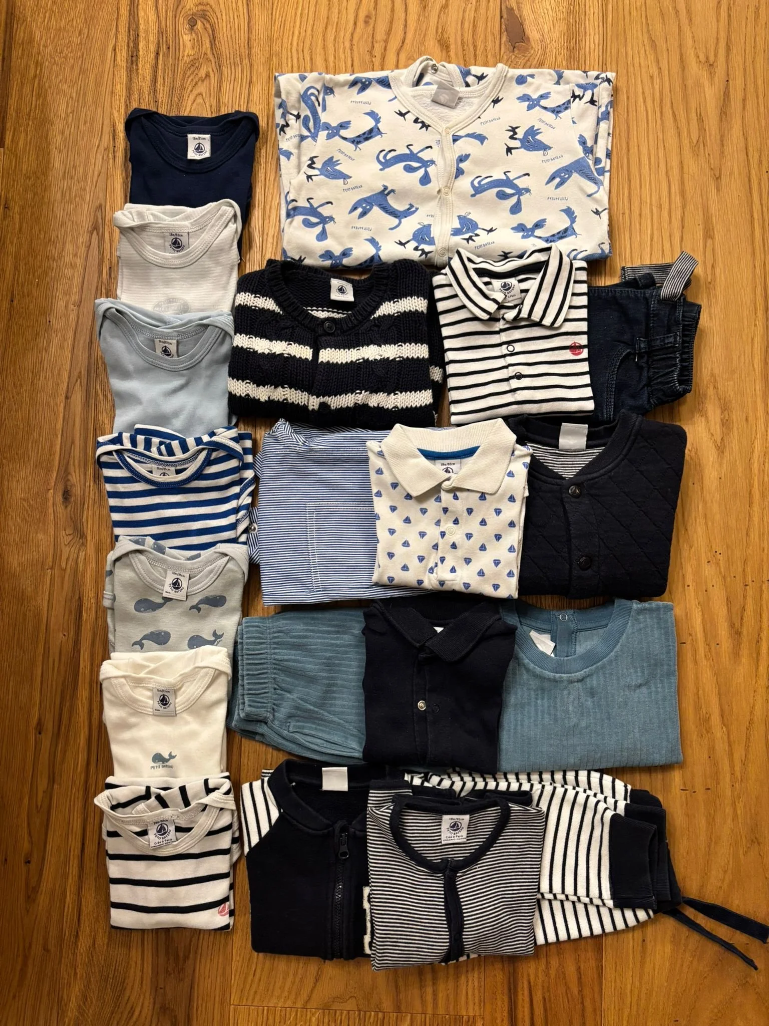 A collection of folded children's clothing arranged on a wooden floor, including shirts, sweaters, and pajamas in various patterns like stripes, whales, and dinosaurs in shades of blue, white, black, and gray.