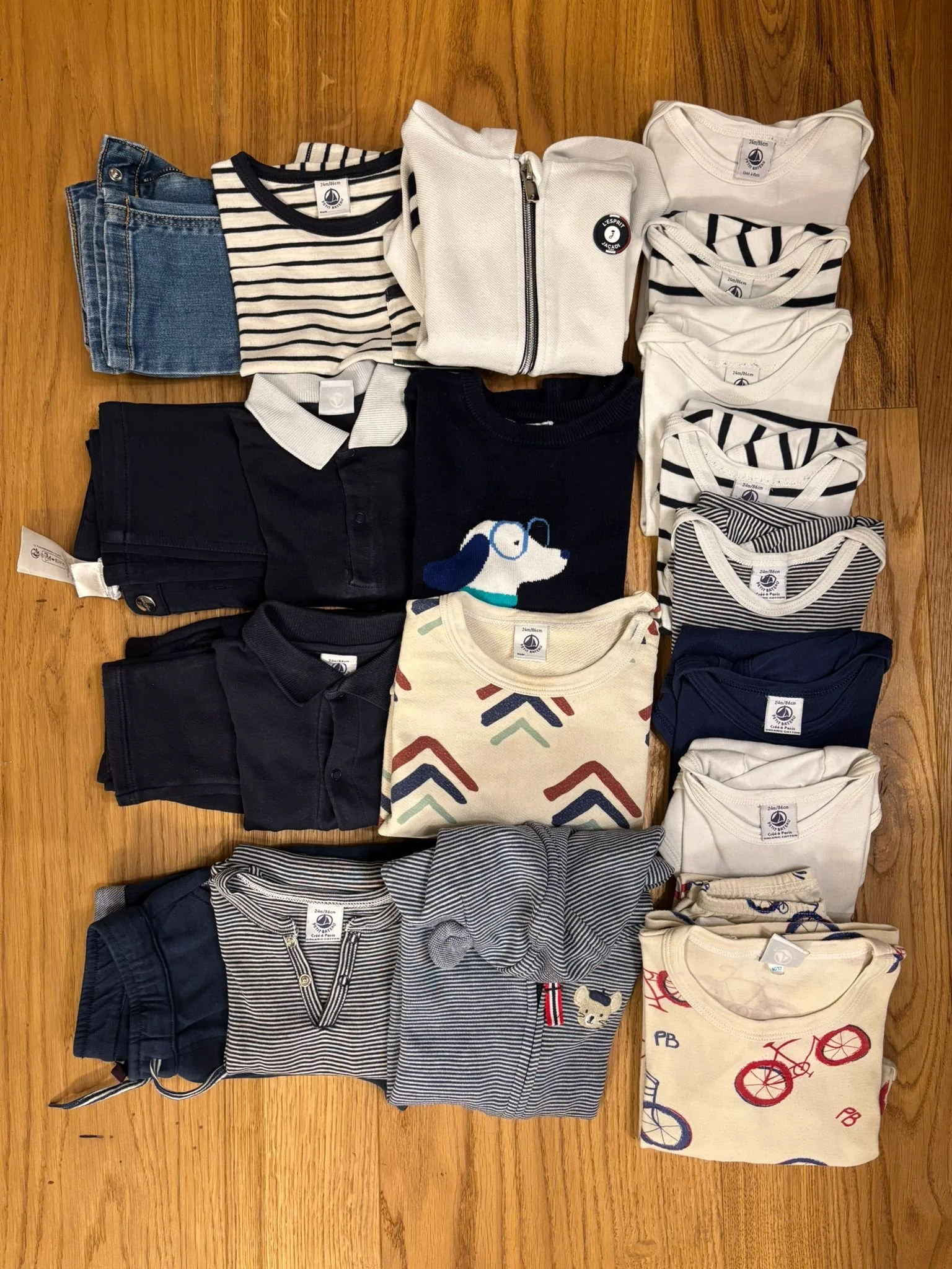 A collection of folded children's clothing including jeans, striped t-shirts, sweaters, polo shirts, and a hoodie arranged on a wooden surface.