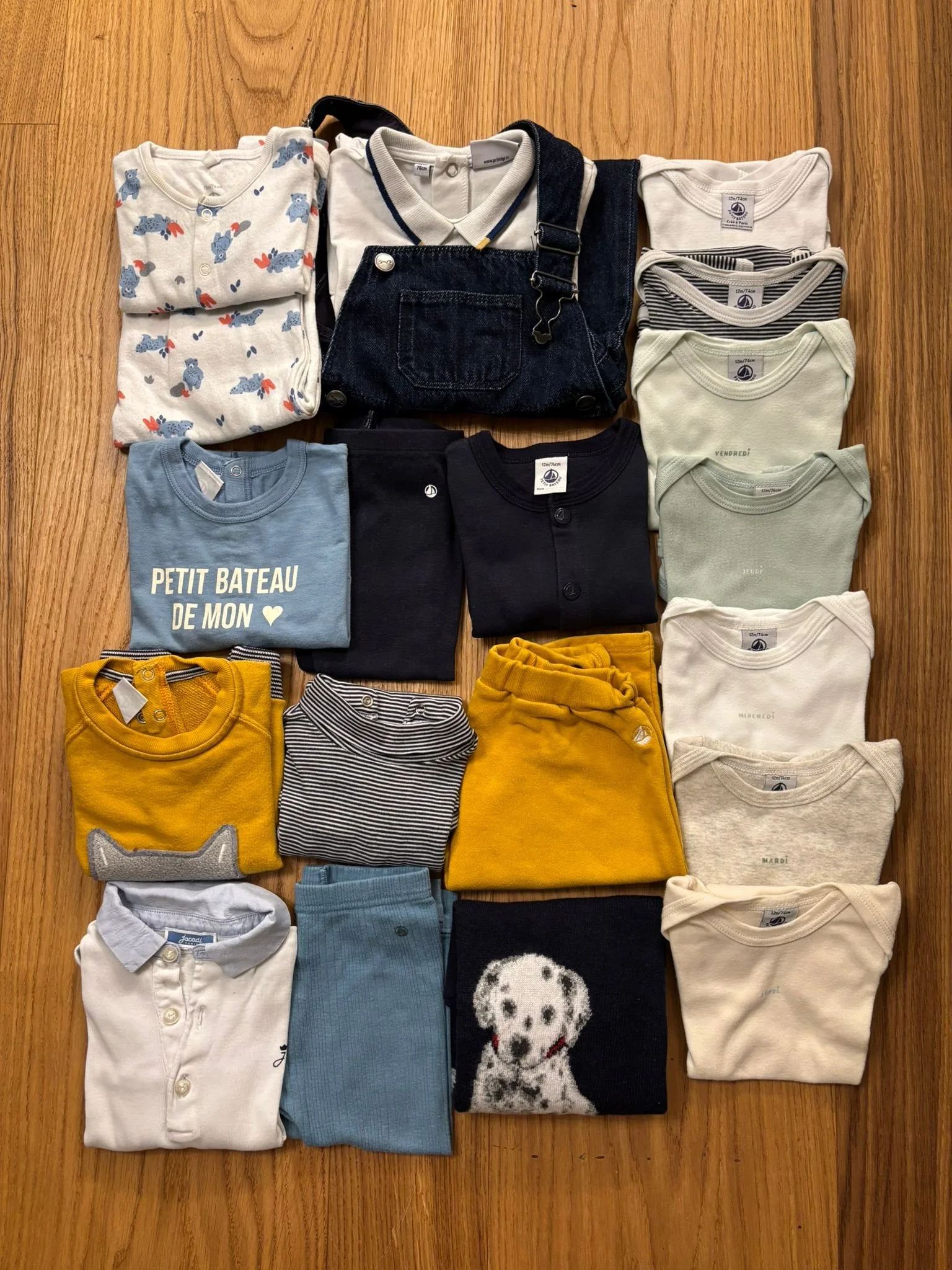 Collection of folded and unfolded children's clothing, including shirts, sweaters, and overalls, arranged on a wooden surface.