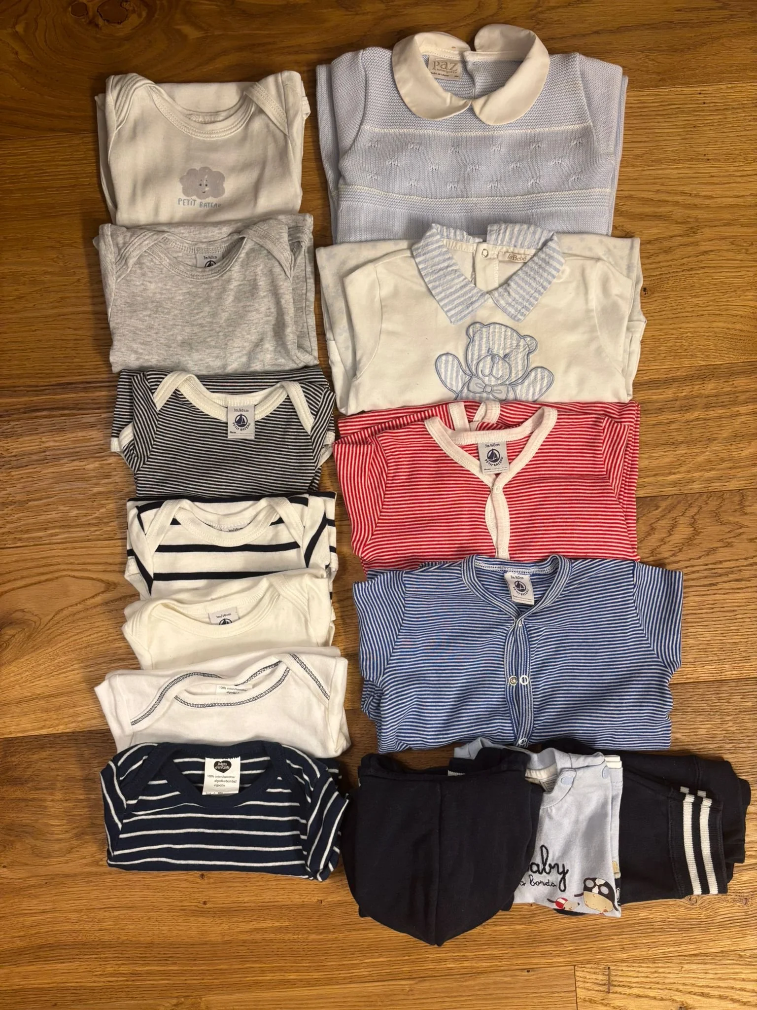 A collection of folded baby clothes on a wooden floor, including onesies, shirts, and sweaters in various colors and patterns such as stripes, solid, and with cartoon characters.
