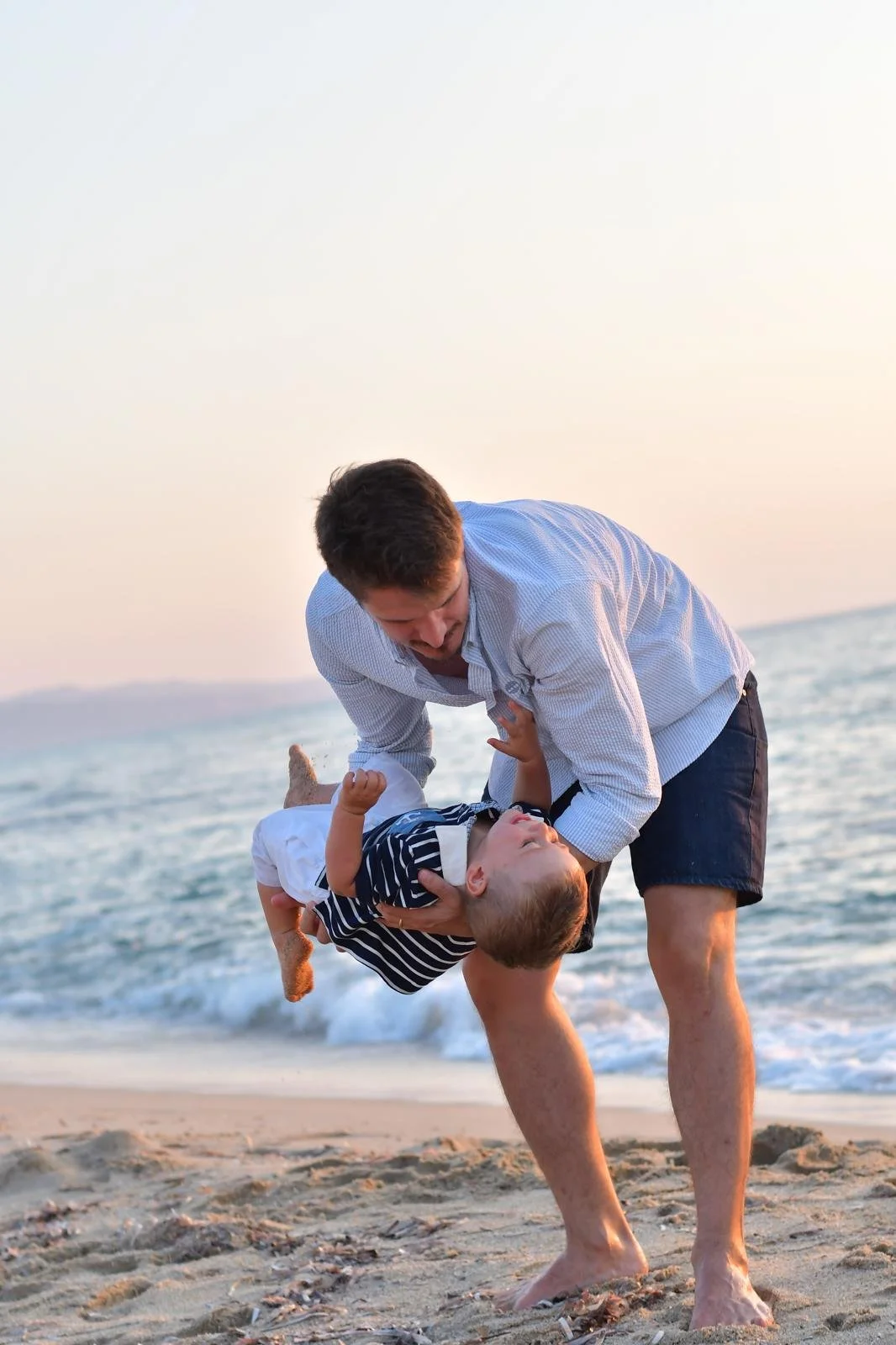 A man playing with a young boy on the beach at sunset, with the man lifting the boy into the air and both smiling, ocean waves in the background.