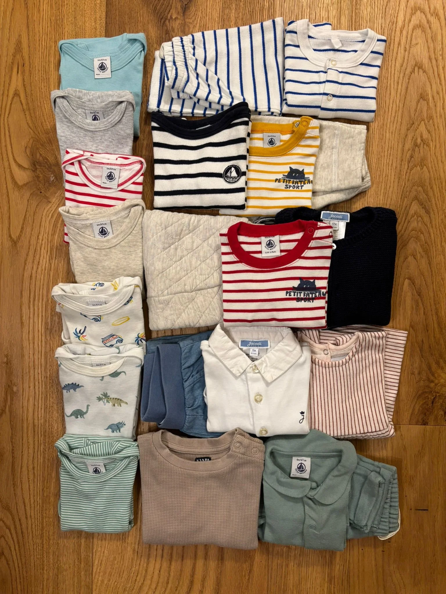 A variety of neatly folded children's clothing items, including striped shirts, solid t-shirts, patterned pajamas, and a quilted layer, arranged on a wooden surface.