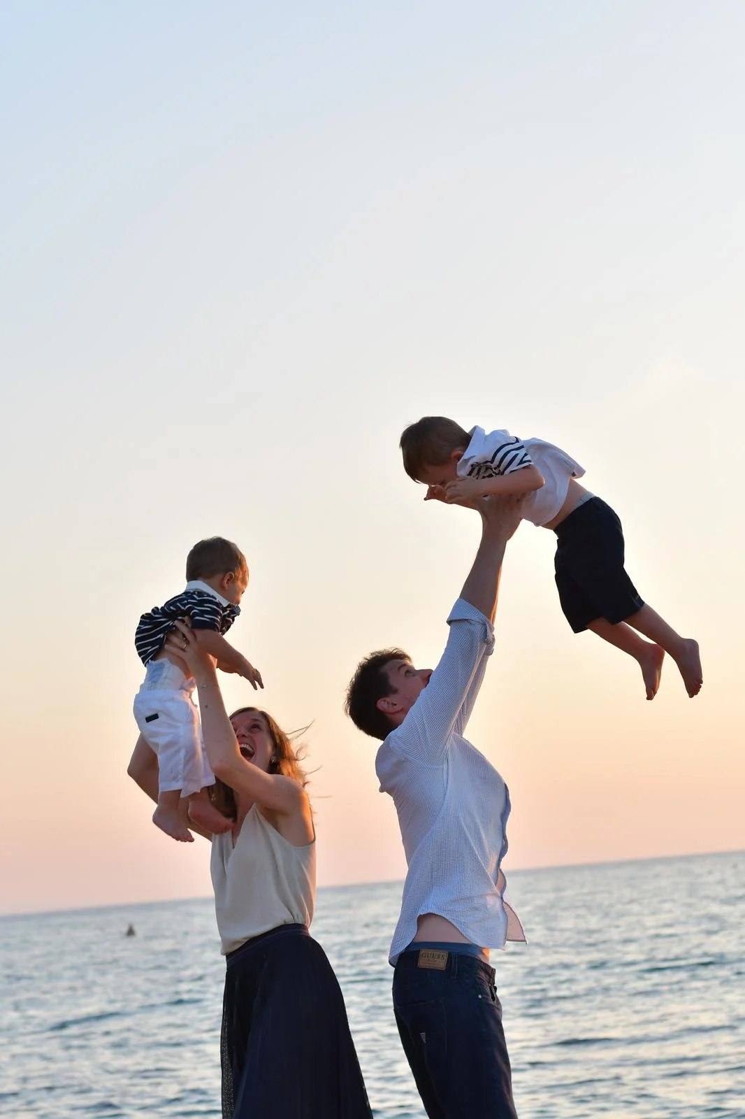 A family of four playing by the water during sunset, with the father tossing a young boy in the air and the mother holding another child, all smiling and enjoying the moment.