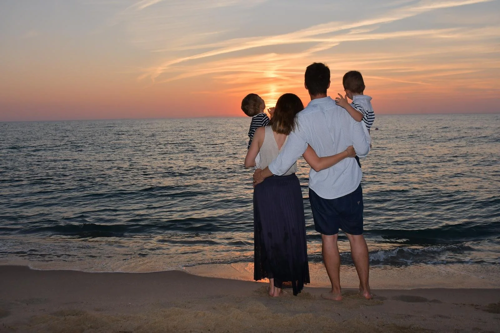 A family of four standing on the beach, watching the sunset over the ocean. The parents are holding their two children, all facing the horizon, with waves gently crashing at their feet.