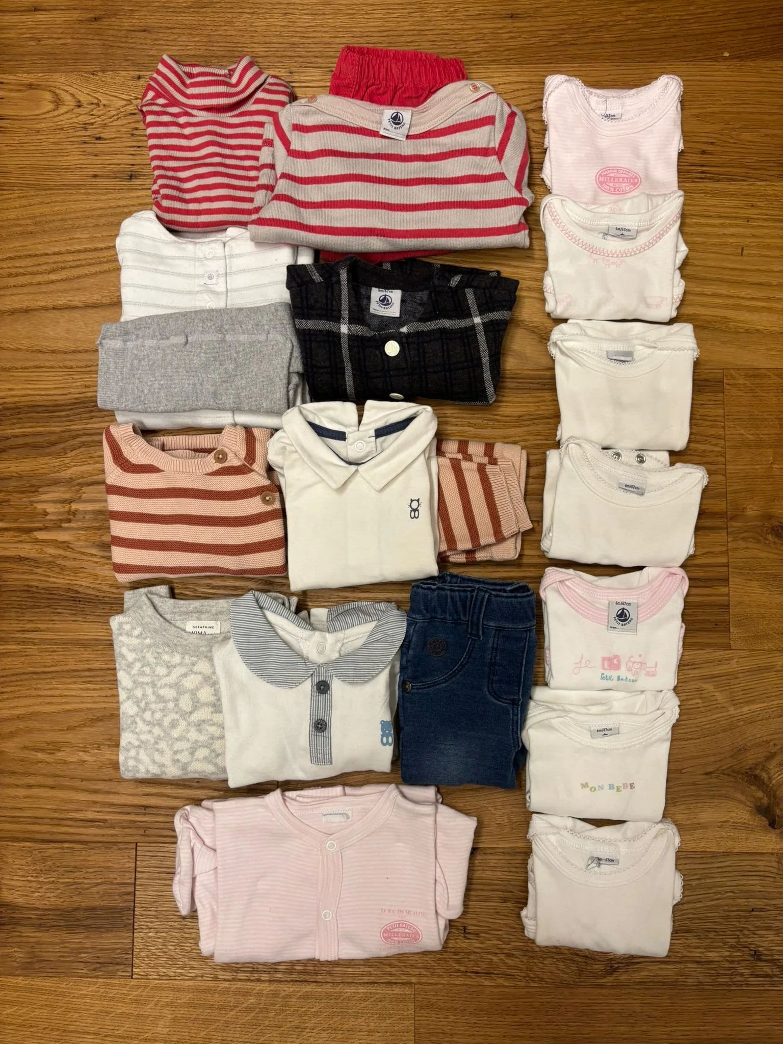 A collection of children's clothing neatly folded and arranged on a wooden floor, including shirts, sweaters, pants, and pajamas in various colors and patterns.