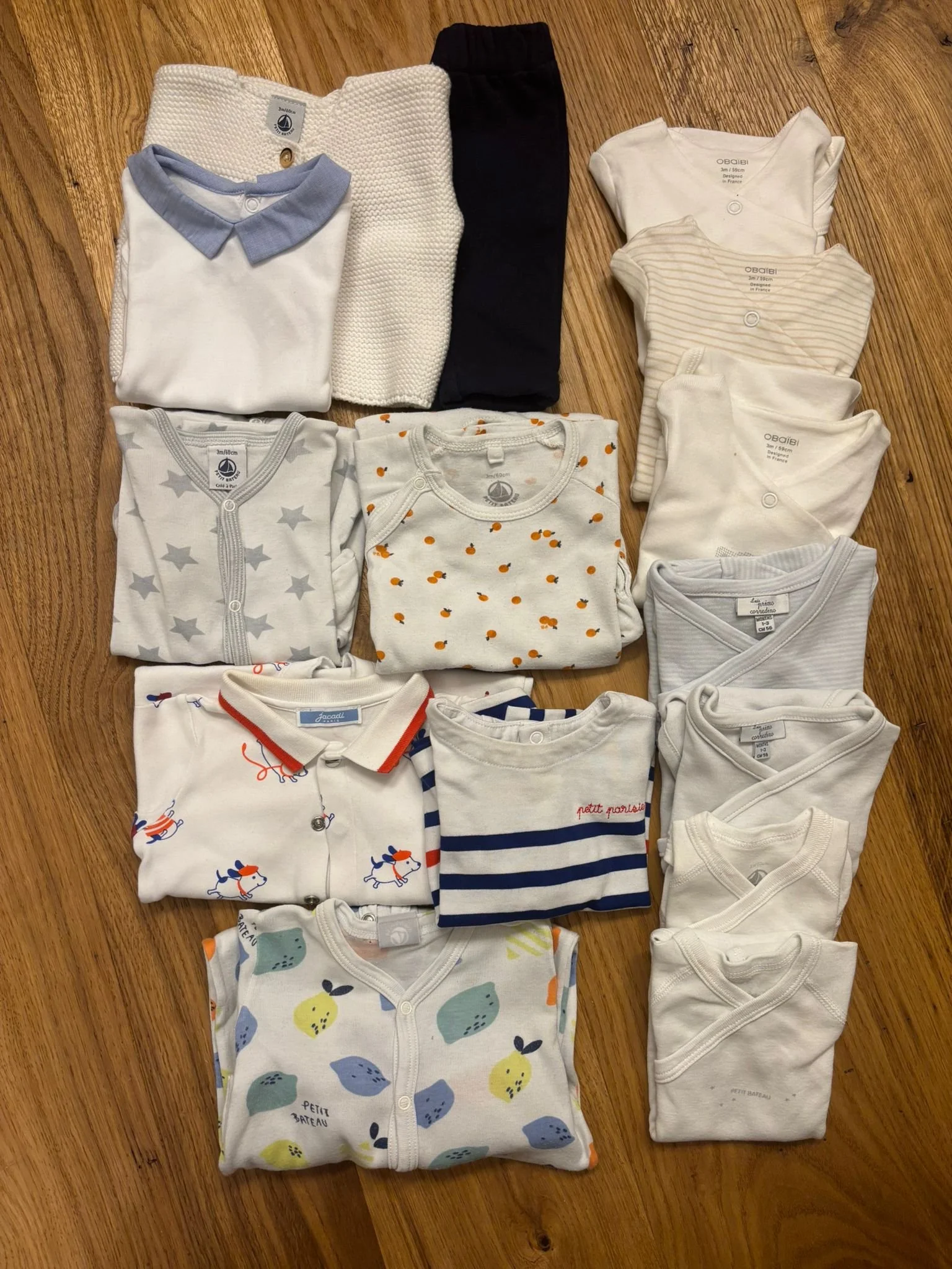 Una serie di vestiti da bambino ordinati su un pavimento di legno, inclusi bodini, maglioni, pantaloni e una camicia con vari disegni e colori.