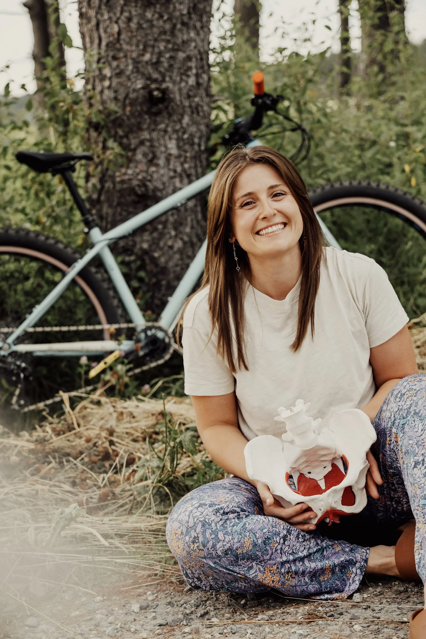 Desirae, the owner of Cadence Pelvic Health and Wellness, sits outdoors on the ground holding a model of a pelvis, with a bicycle leaning against a tree behind her, in a natural forest setting.