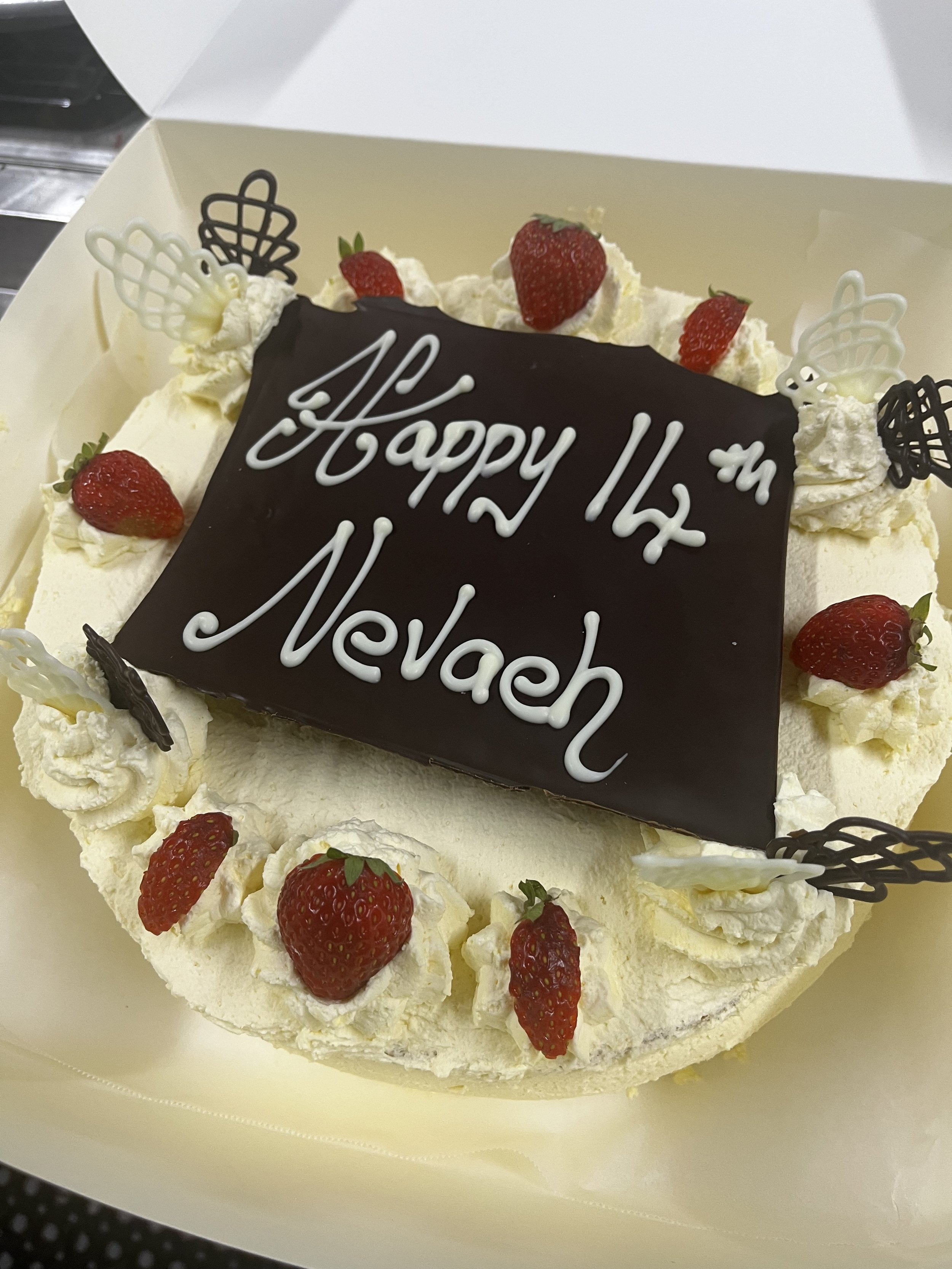 A birthday cake decorated with strawberries, whipped cream, and chocolate pieces in a white box, with a chocolate plaque that says 'Happy 14th Melwach'.