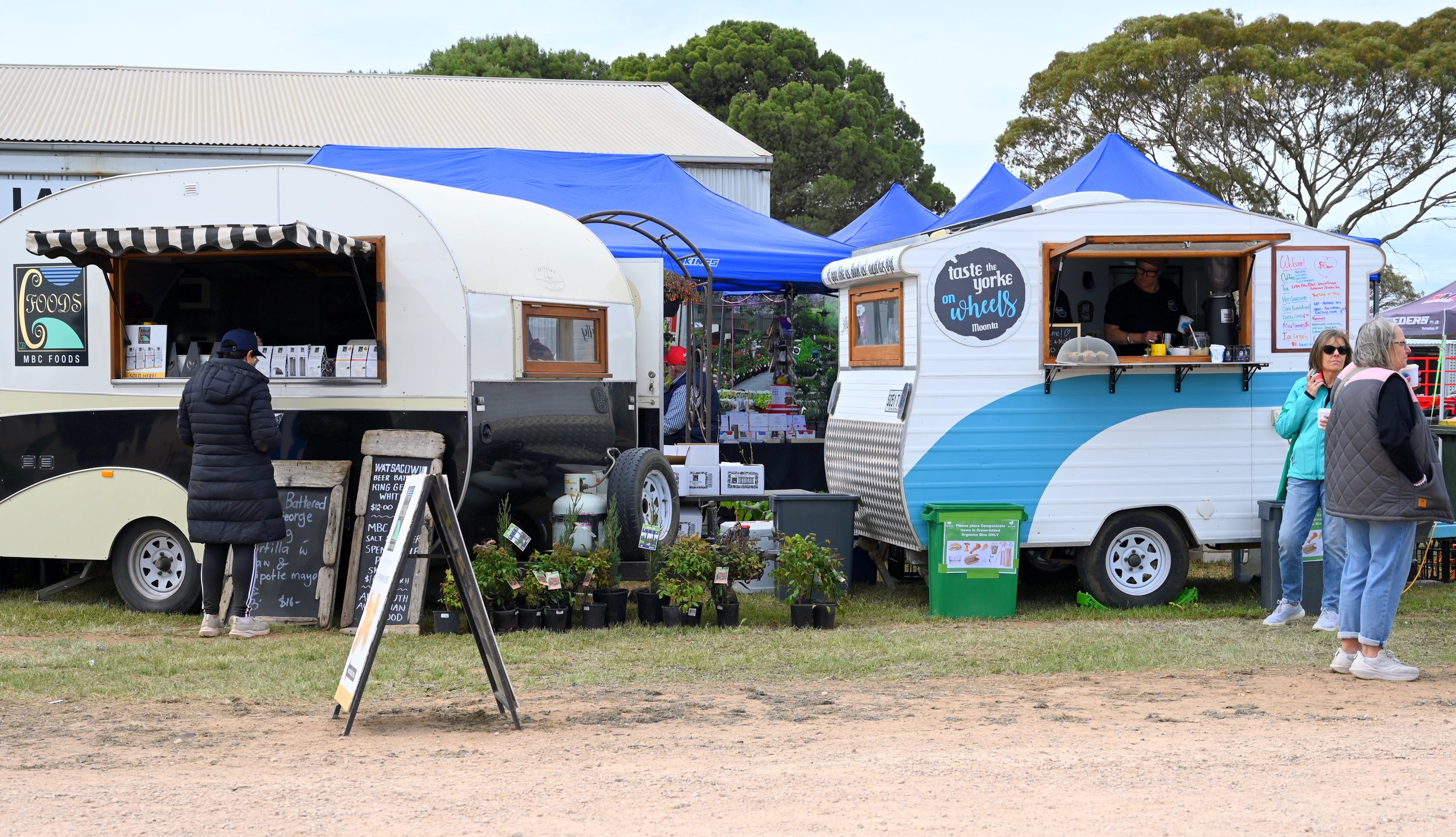 Two food trucks parked side by side at an outdoor event, with people standing nearby. The left truck has a black and white sign reading 'FOODS,' and the right truck has a sign 'taste the yorke on wheels.' Potted plants and a green trash bin are in fr