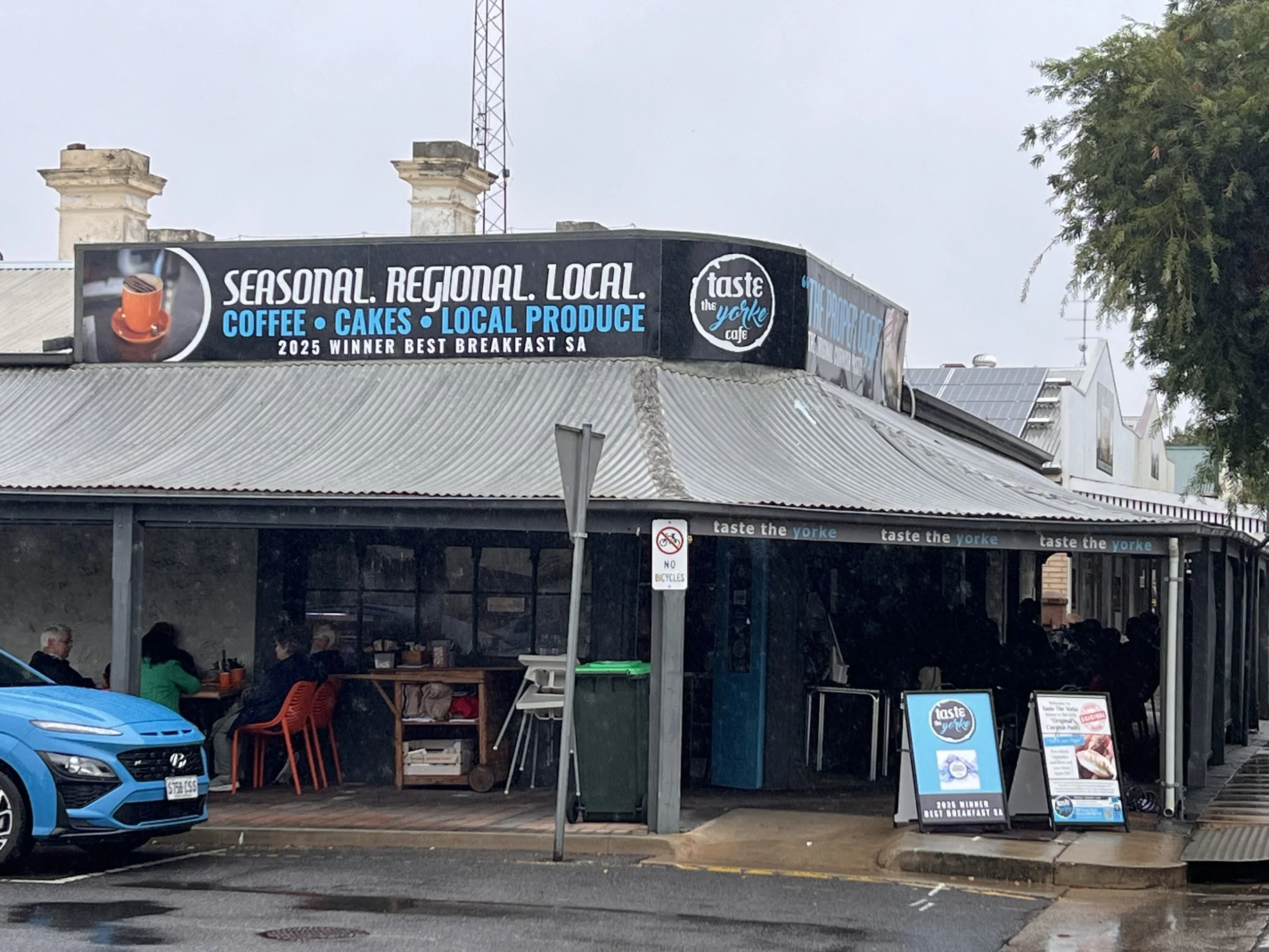 A cafe sign advertising seasonal, regional, and local coffee, cakes, and produce, with outdoor seating and a few people sitting underneath a roof.