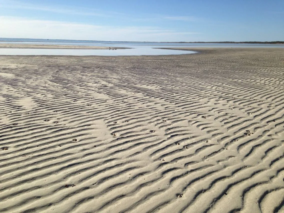 Calm beach with rippled sand leading to water, shoreline, and blue sky.