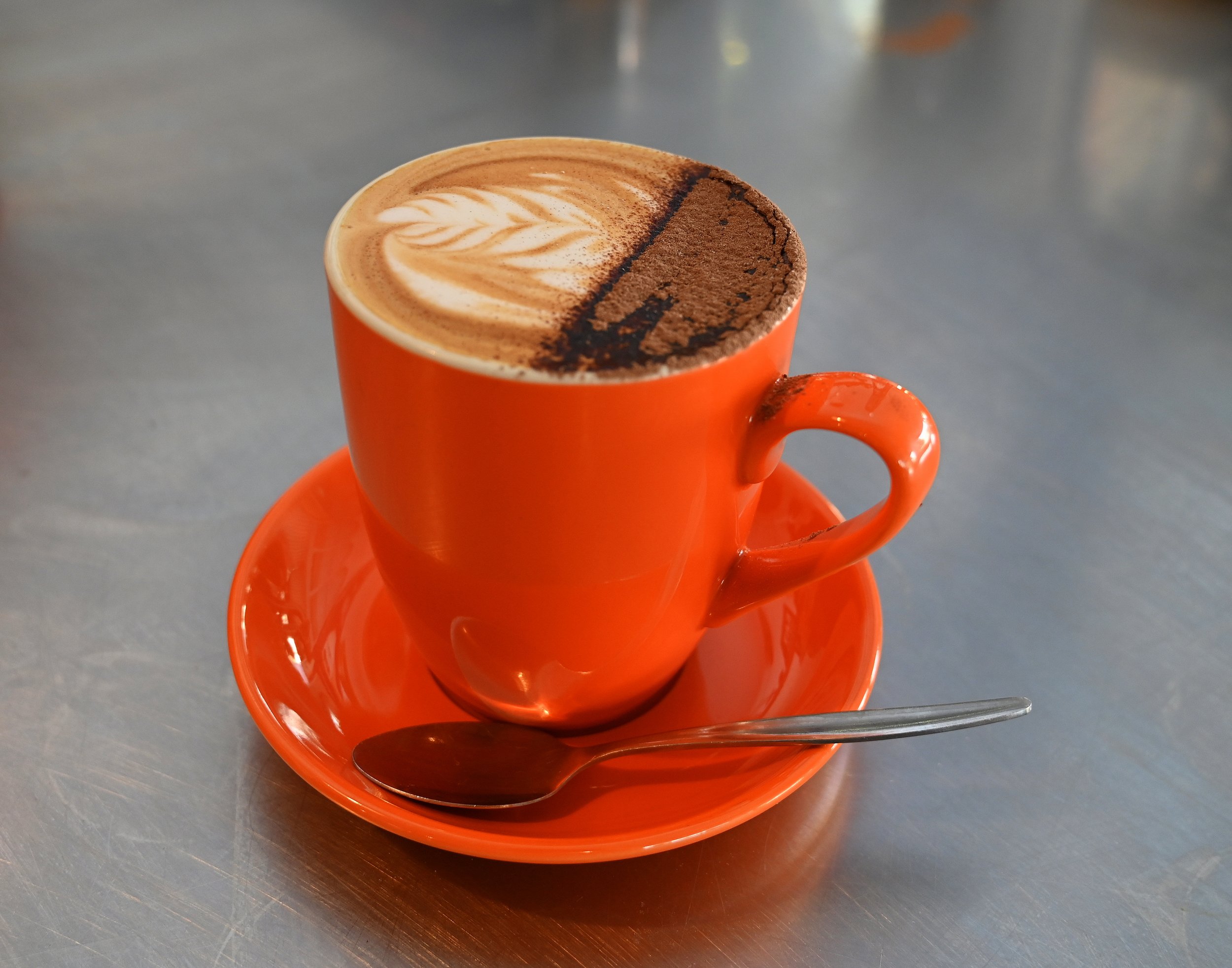A frothy coffee beverage with latte art in an orange cup and saucer, accompanied by a metallic spoon, on a metallic table.