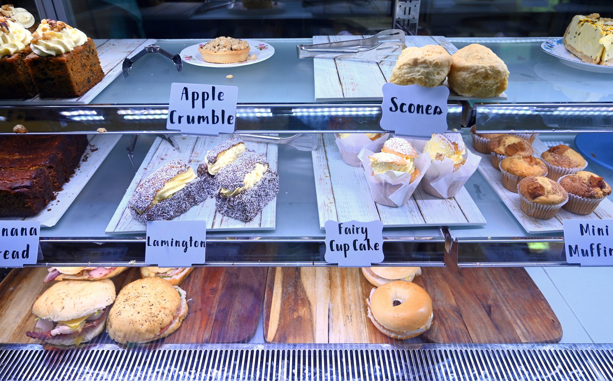 A display case with various baked goods including apple crumble, scones, banana bread, lamingtons, fairy cup cakes, mini muffins, sandwiches, and a bagel, each labeled with handwritten signs.
