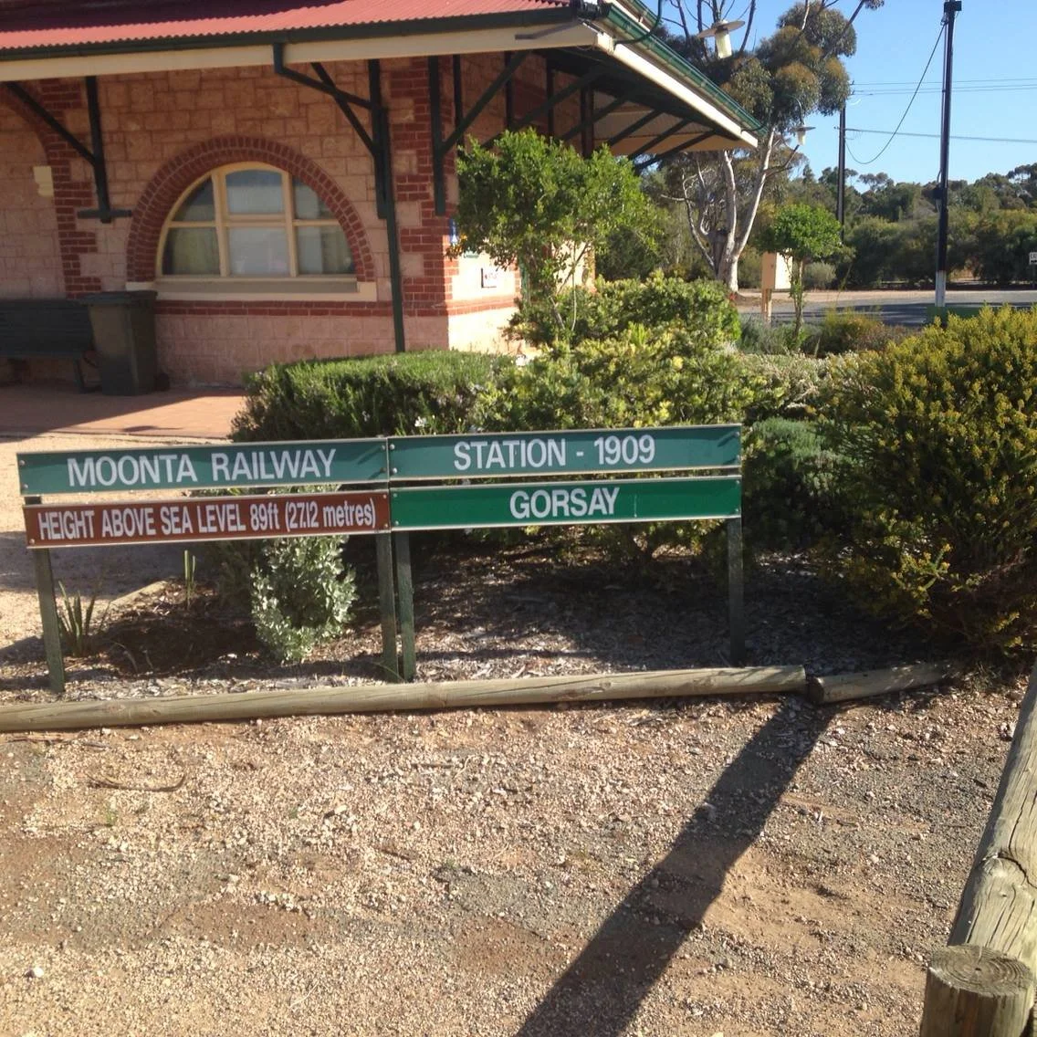 Signpost at Moonta Railway Station 1909 indicating height above sea level at 89 feet or 27.12 meters, with a brick building and greenery in the background. Moonta Mines Railway