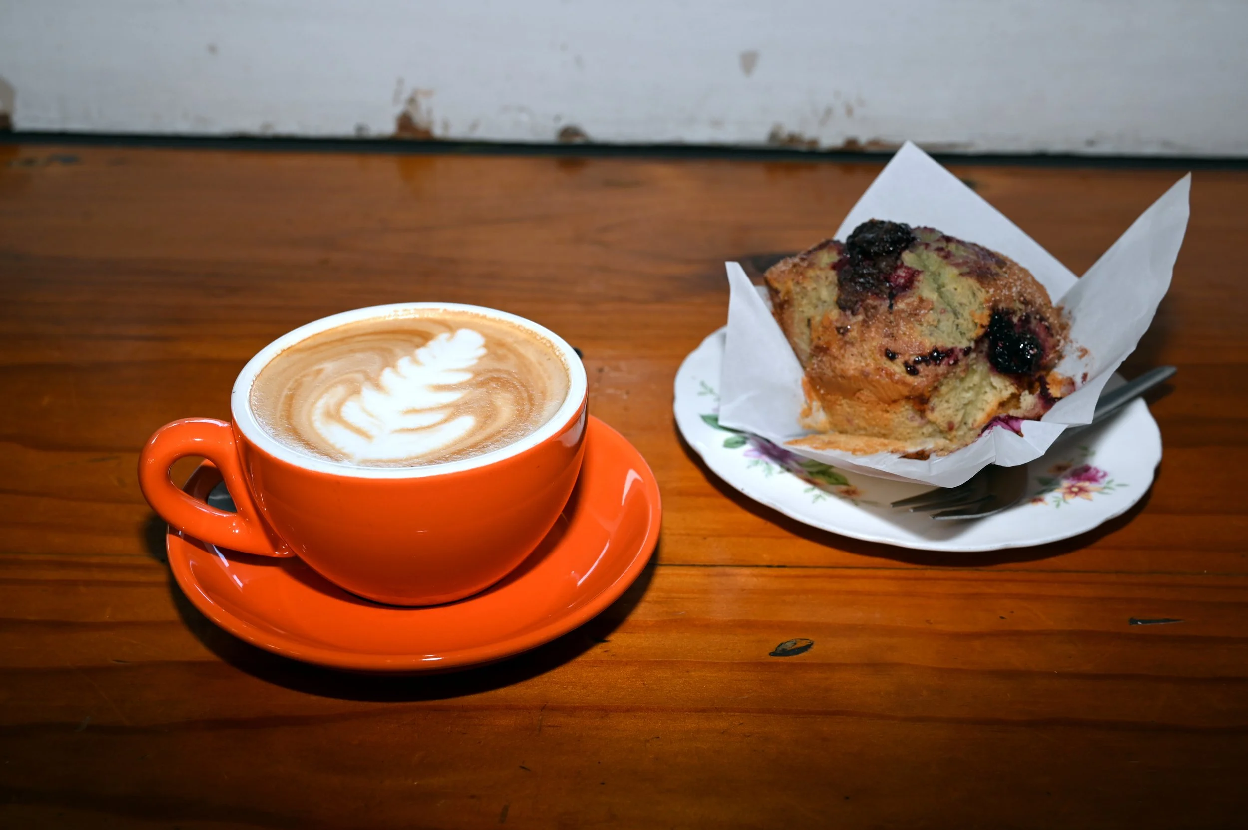 A cup of coffee with latte art on a saucer and a blueberry muffin on a floral plate on a wooden table.