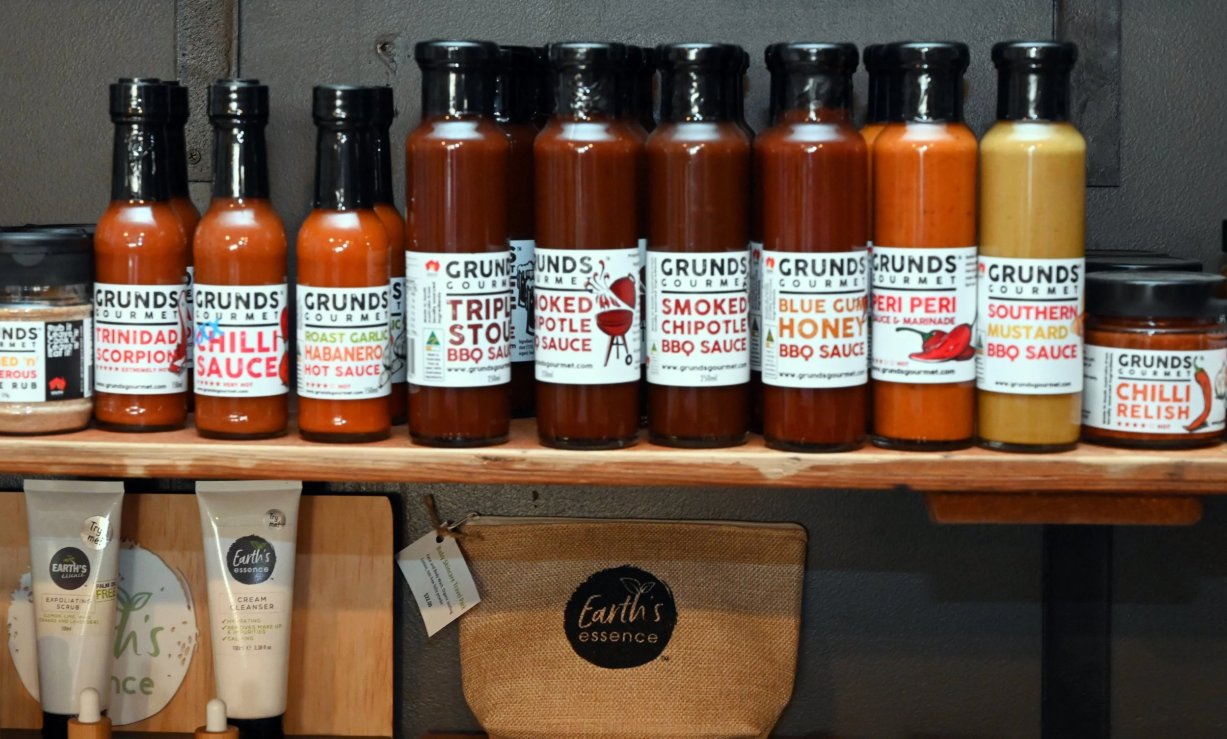 Shelf displaying various bottles of BBQ sauces with brand labels, including Trinidad Scorpion, Chilli Sauce, Roasted Garlic Habanero Hot Sauce, Smoked Tpotle BBQ Sauce, Smoked Chipotle BBQ Sauce, Blue Cumin Honey BBQ Sauce, Peri Peri Sauce, Southern 