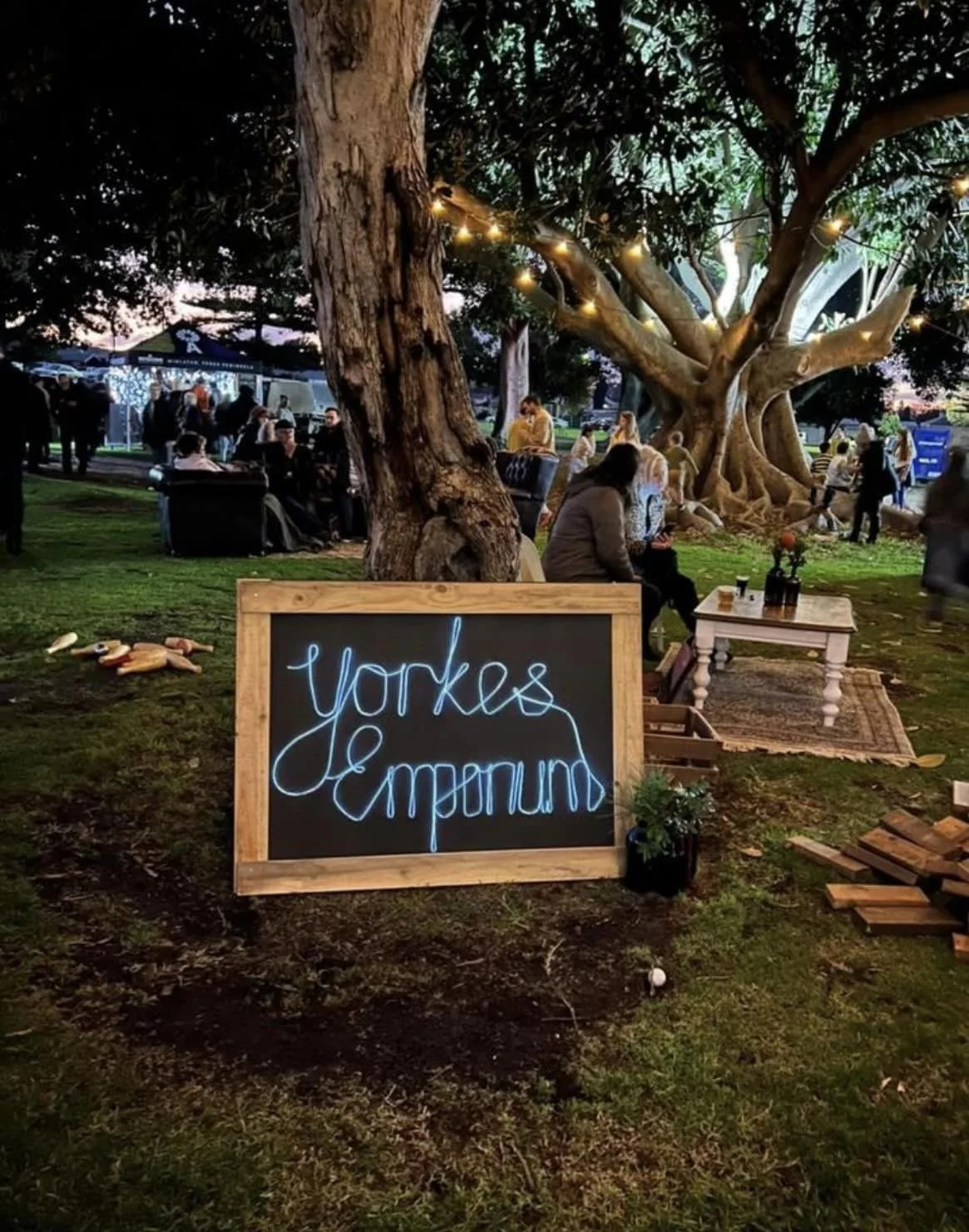 A scene of an outdoor event or gathering during the evening with people sitting and walking near a large tree decorated with string lights. In the foreground, there is a blackboard sign with glowing blue neon-style writing that says 'Yorges Empound',