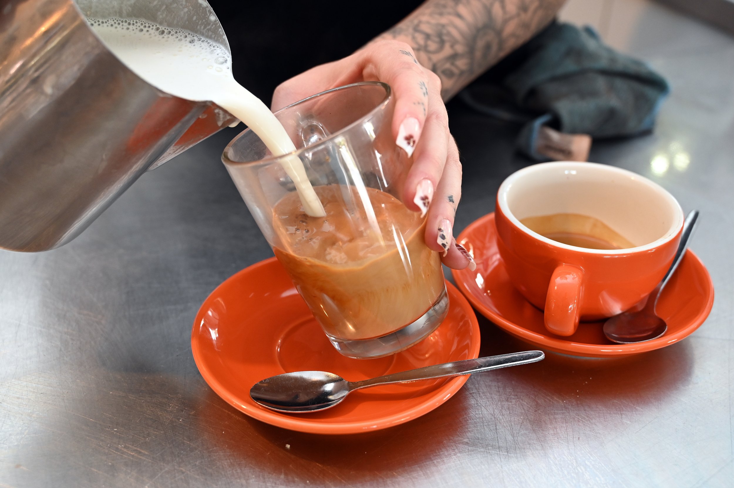 Pouring cream into a glass of iced coffee on an orange saucer, with a cup of hot coffee on an orange saucer nearby, on a metal table.