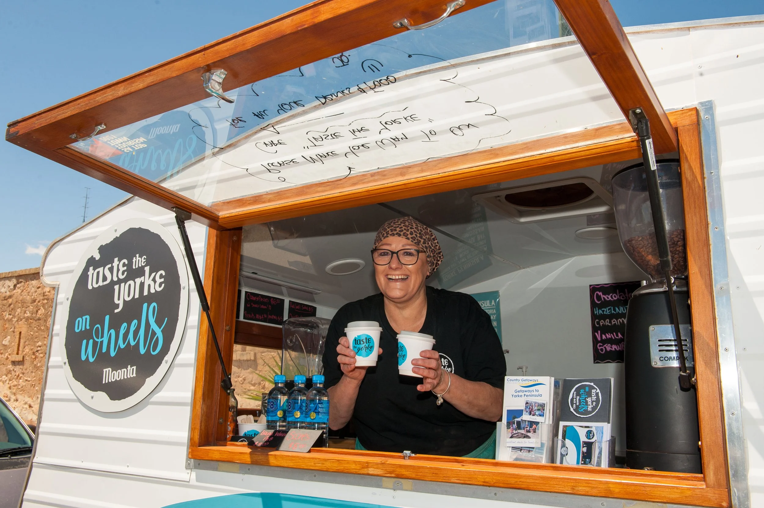 A smiling woman with glasses and a leopard print headscarf serving drinks from a mobile food truck named "Taste the Yorke on Wheels" in Moonta, Australia.