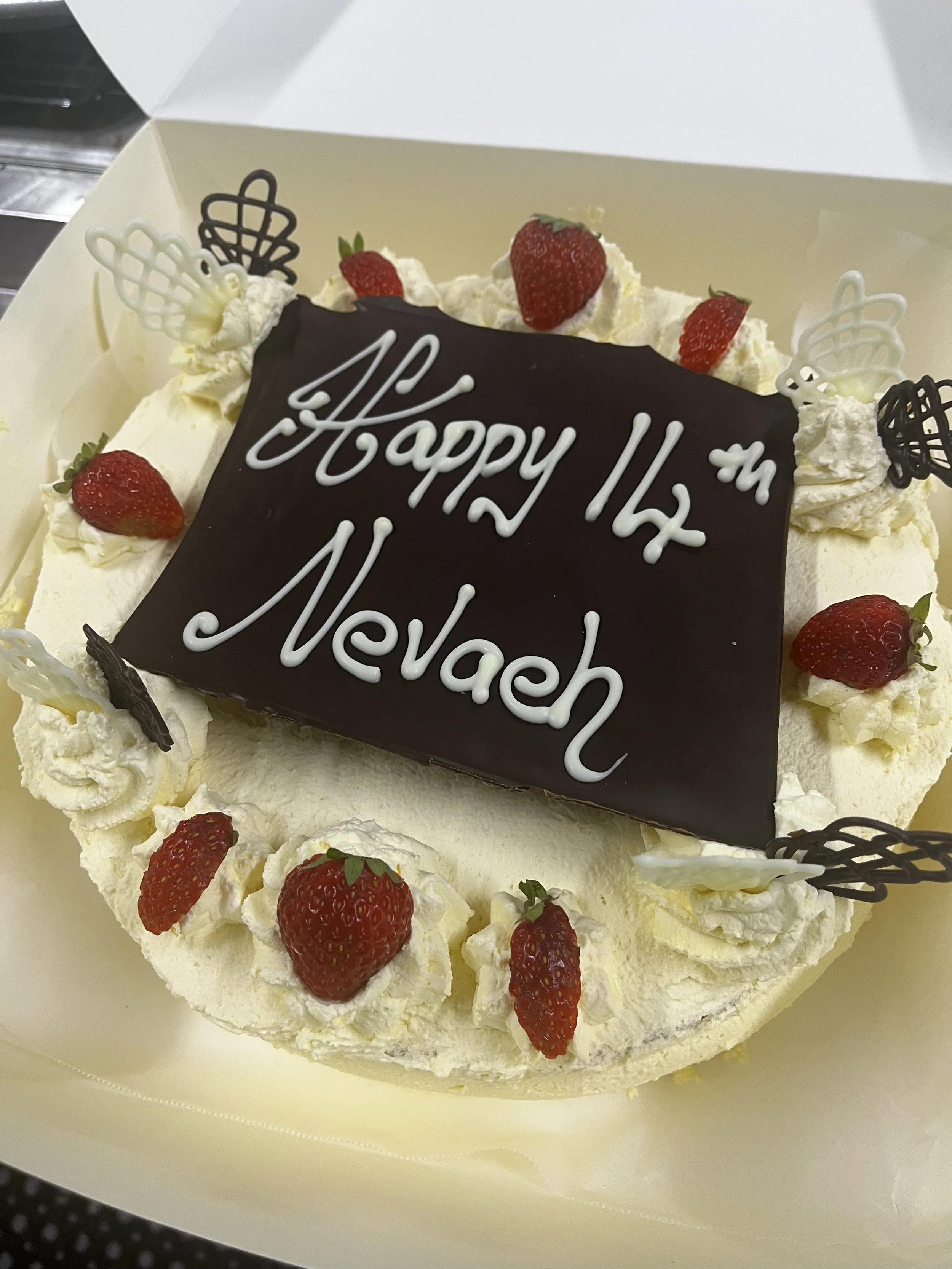 A birthday cake with strawberries, whipped cream, and chocolate decorations, with a chocolate plaque that says 'Happy 14th'. in a white cake box.