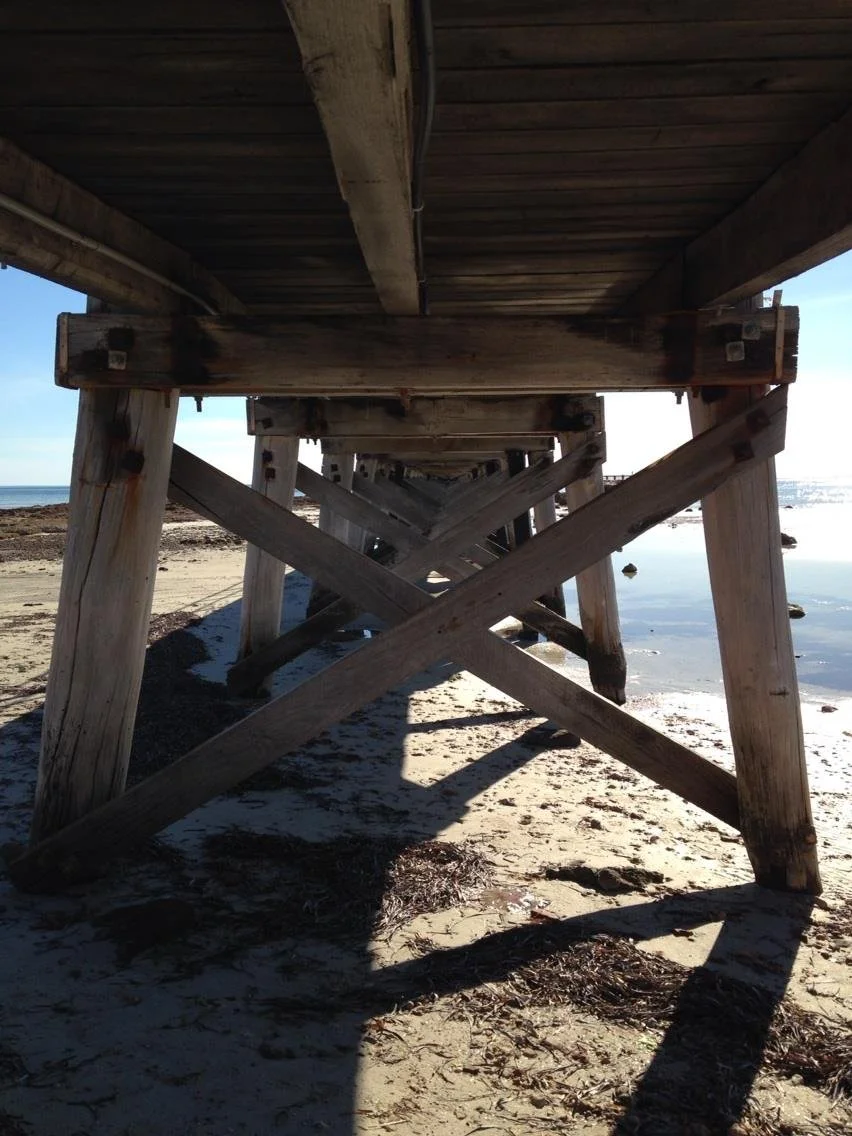 View from underneath Moonta Bay jetty extending over a sandy beach and water, with shadows cast on the sand.