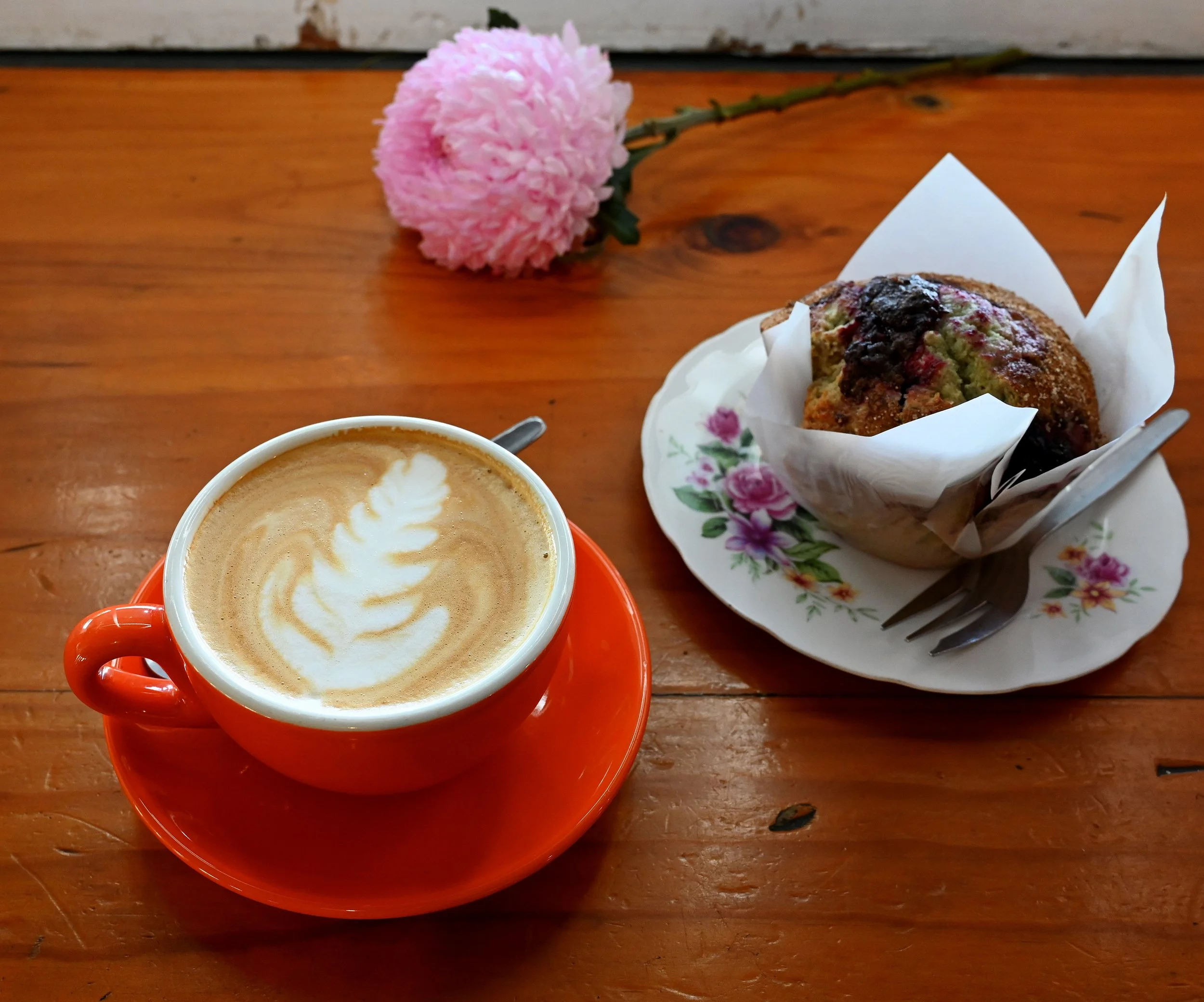 A cup of coffee with latte art in a red cup and saucer, a blueberry muffin on a floral plate with a fork, and a pink carnation flower on a wooden table.
