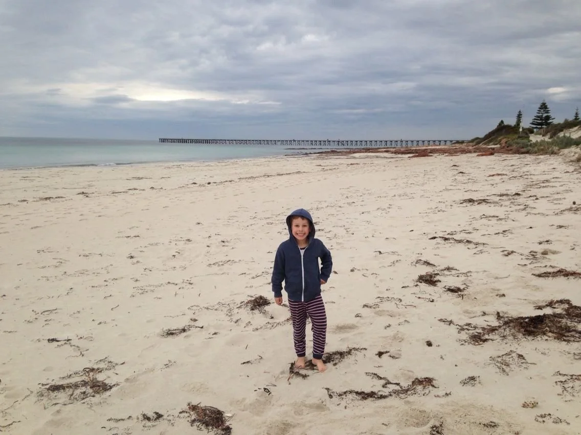 A young child standing barefoot on a sandy beach, wearing a dark hoodie and striped pants, smiling at the camera with overcast skies and a pier in the distance.